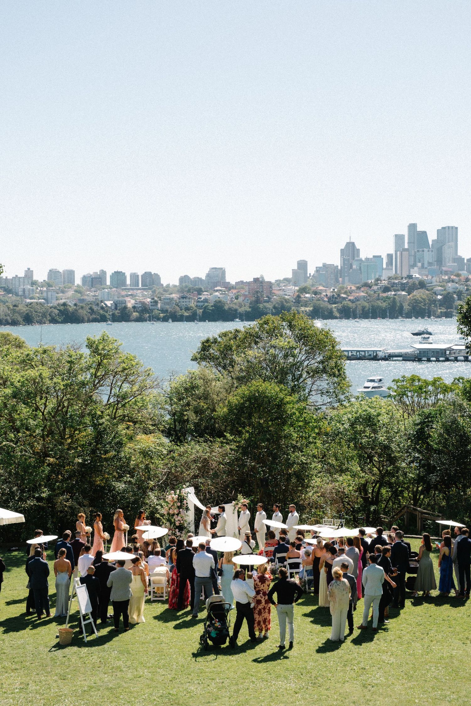 Outdoor wedding ceremony on a lawn overlooking Sydney Harbour with city skyline, guests gathered under white parasols.