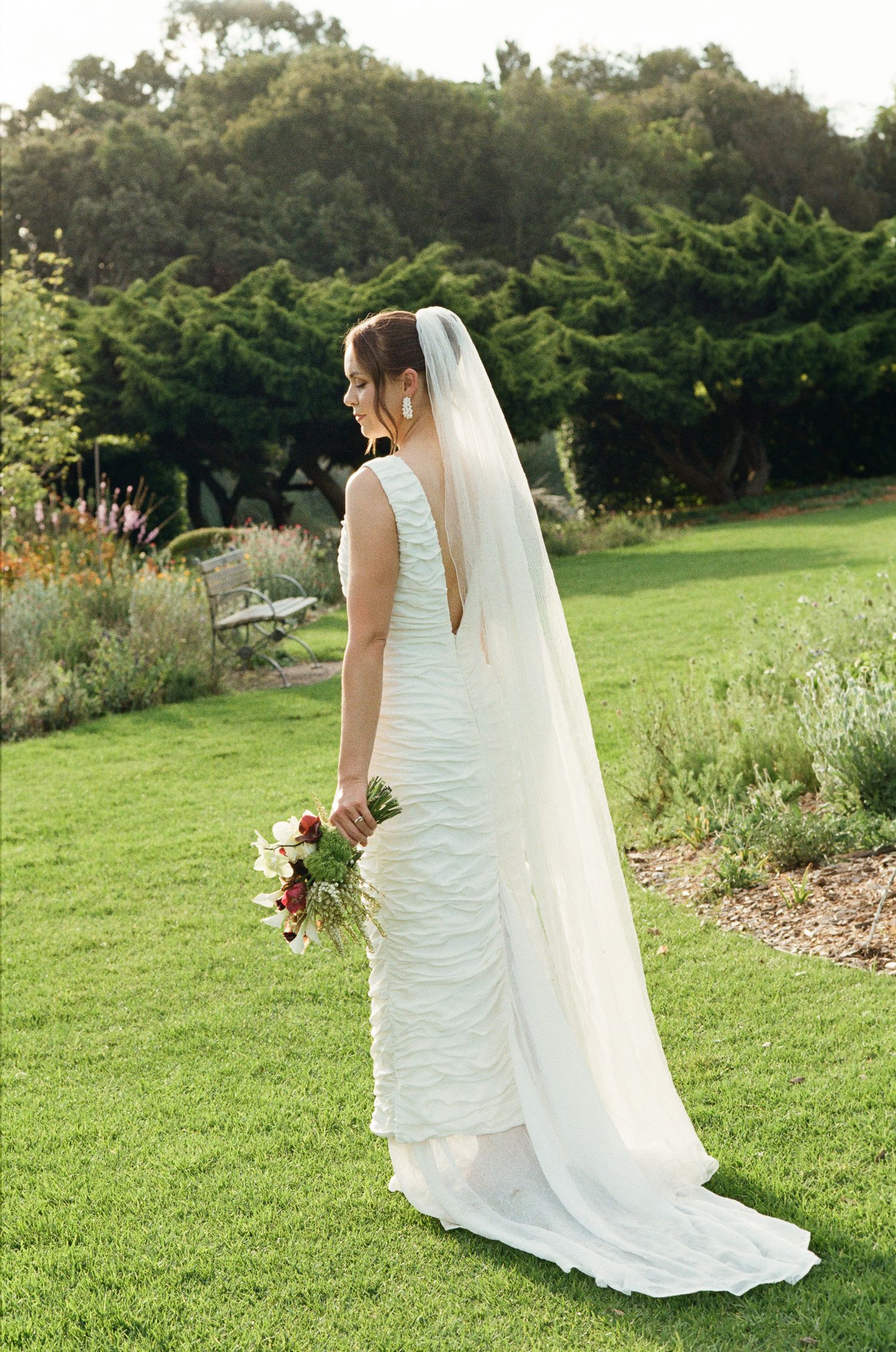 Bride in a fitted ruched white gown and long veil holds a red and white bouquet while standing on a sunlit garden lawn.