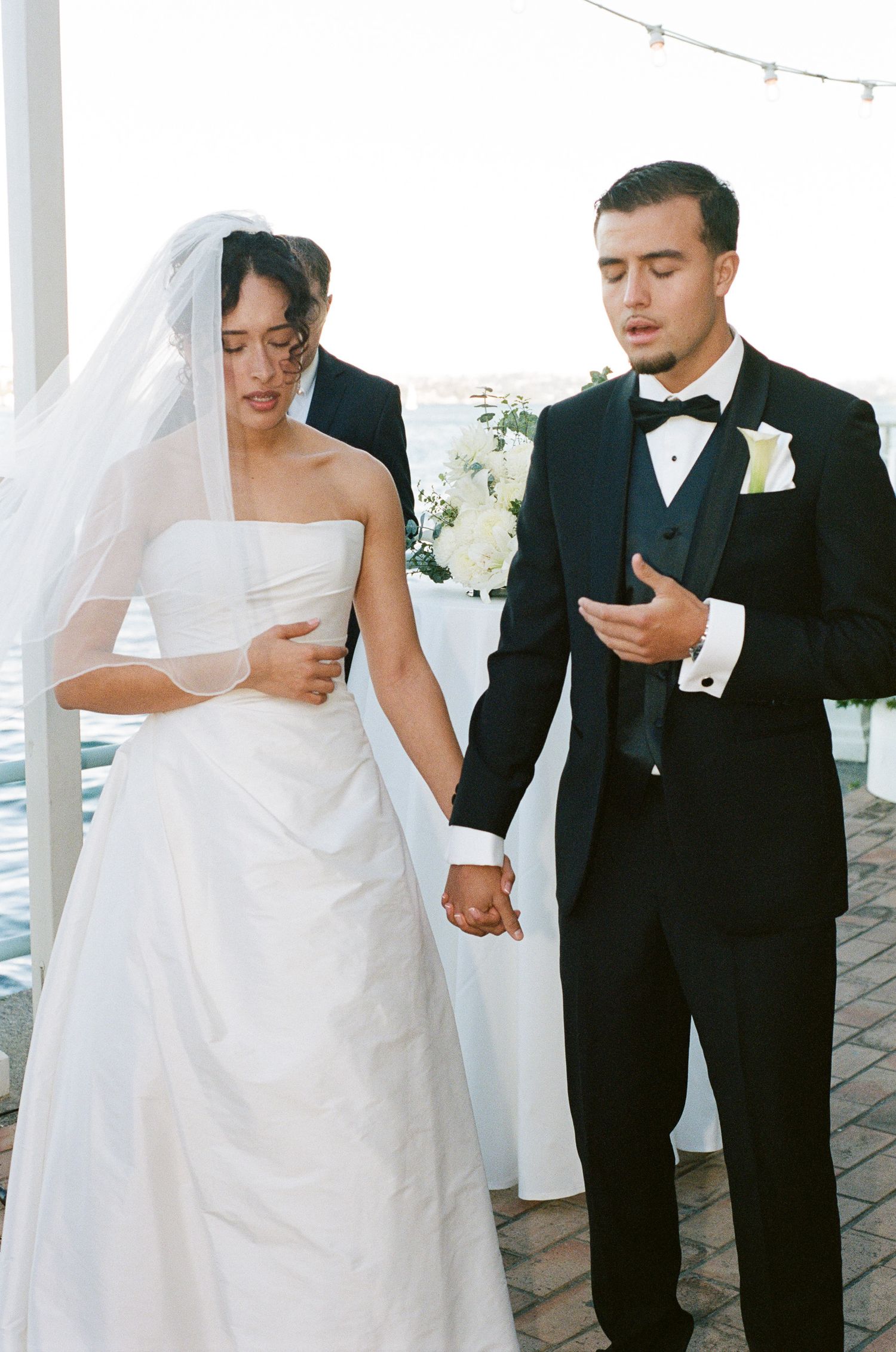 Bride in a strapless white gown and veil holds hands with a groom in a black tuxedo and bow tie during a waterfront wedding ceremony.
