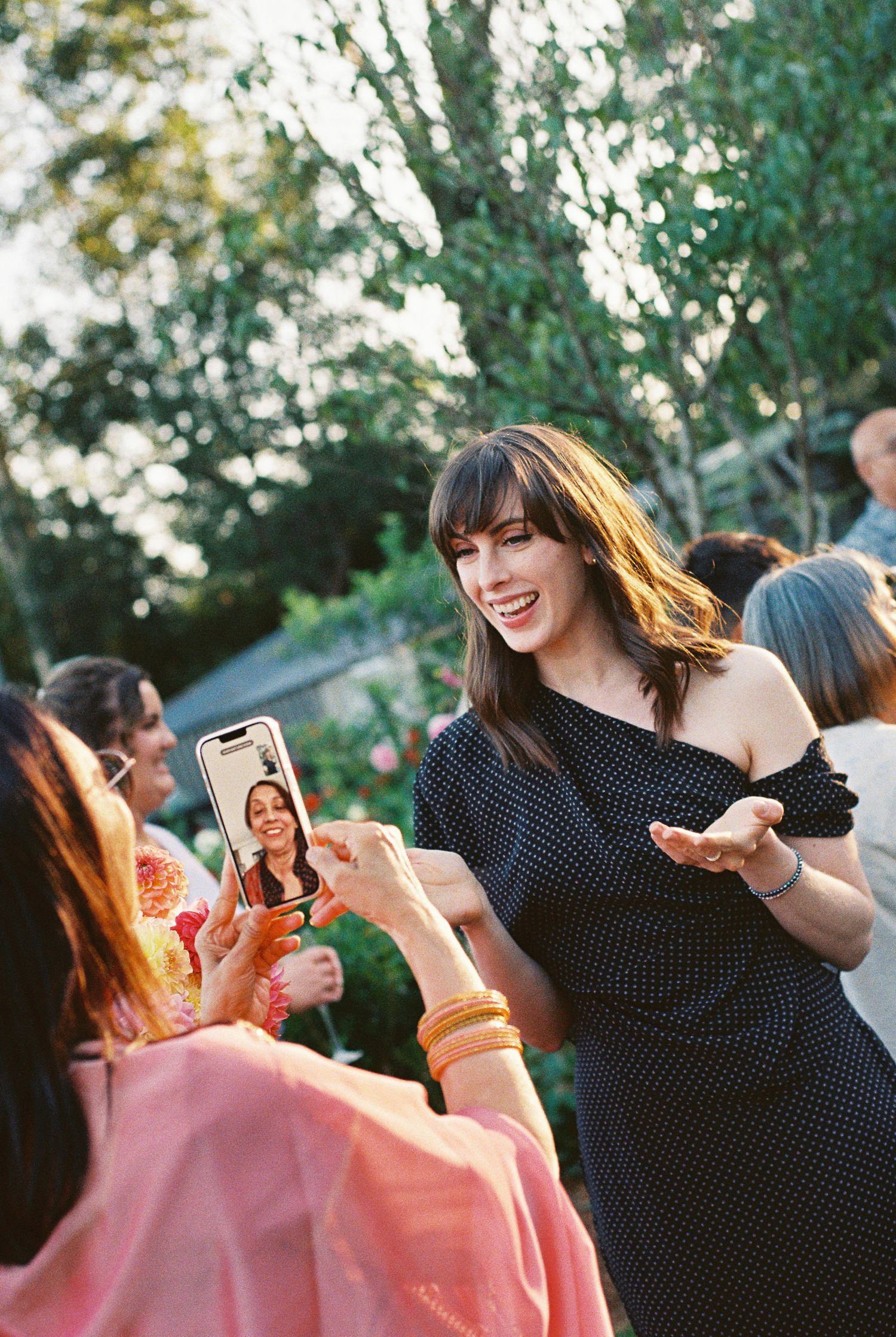 Smiling woman in a black polka dot dress gestures happily at a smartphone video call held by a guest at an outdoor garden party.
