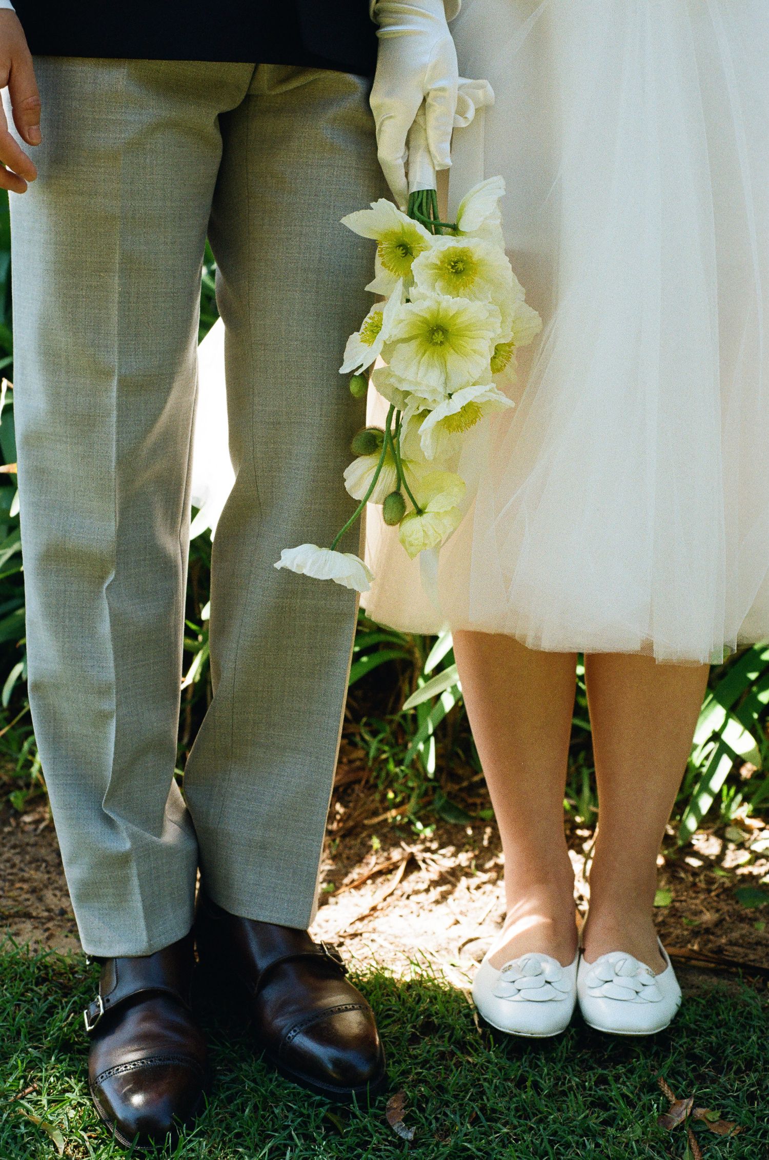 Bride in white flats and tulle skirt holds a bouquet of white flowers beside a groom in gray trousers and brown shoes on grass.