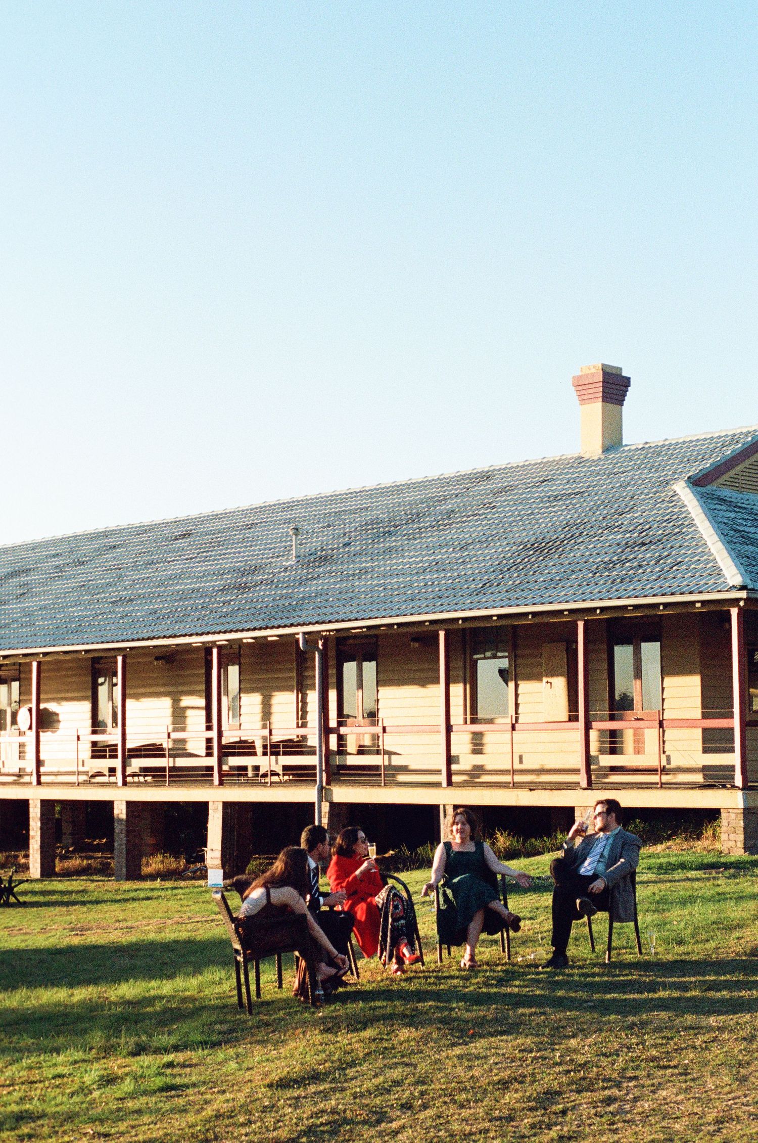 Wedding guests relax on chairs outside a rustic wooden venue building in golden evening light.