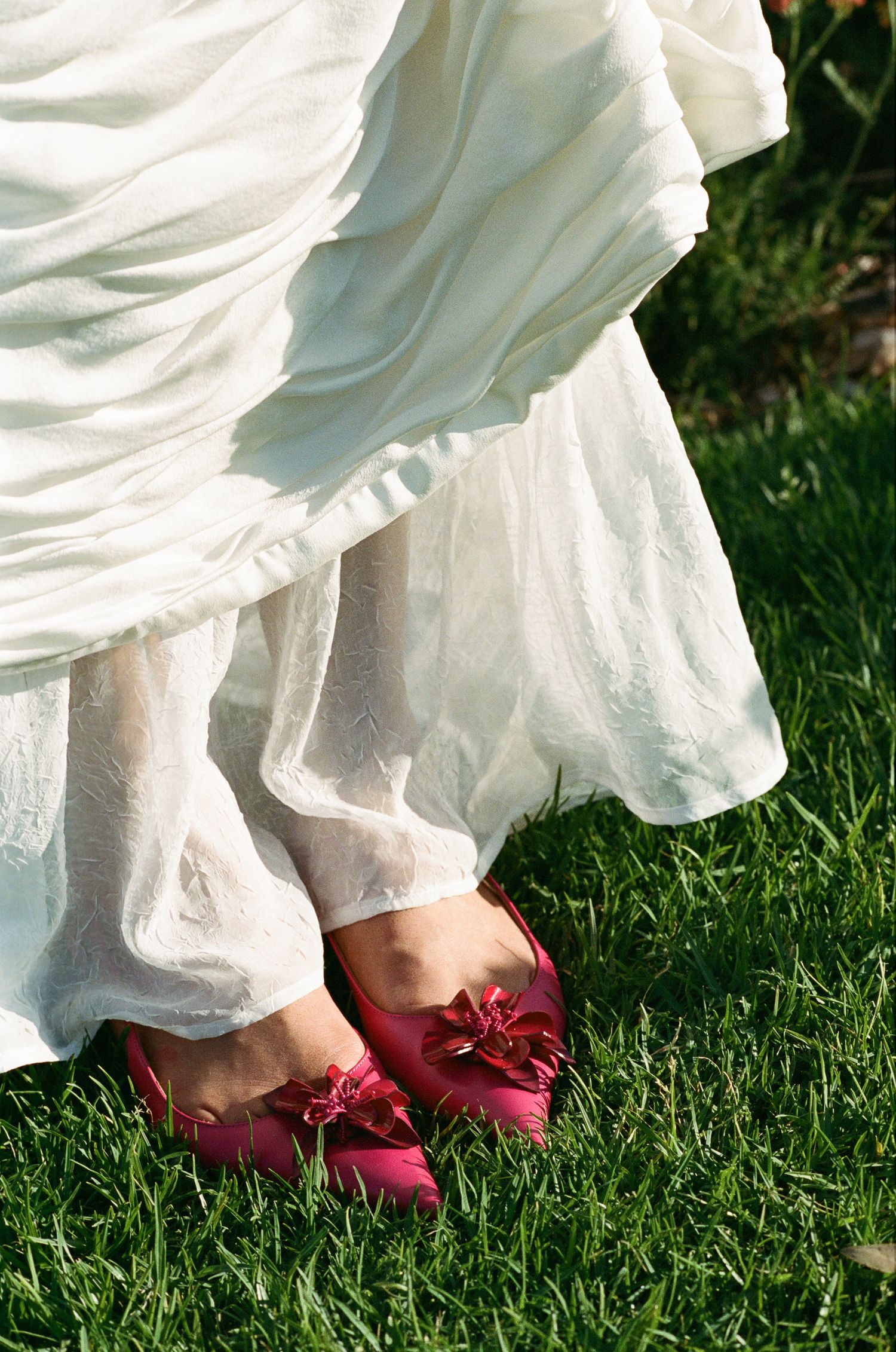 Bride's feet in pink satin flats with bow details standing on green grass beneath a flowing white wedding dress.