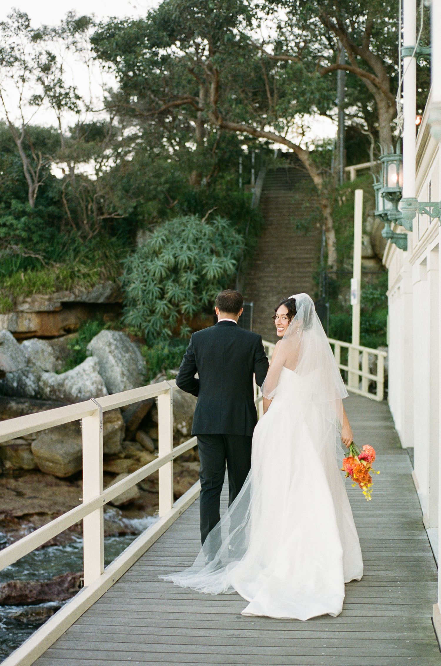 Bride in a white gown with a long veil and groom in a black suit walk along a waterfront wooden boardwalk, holding an orange bouquet.