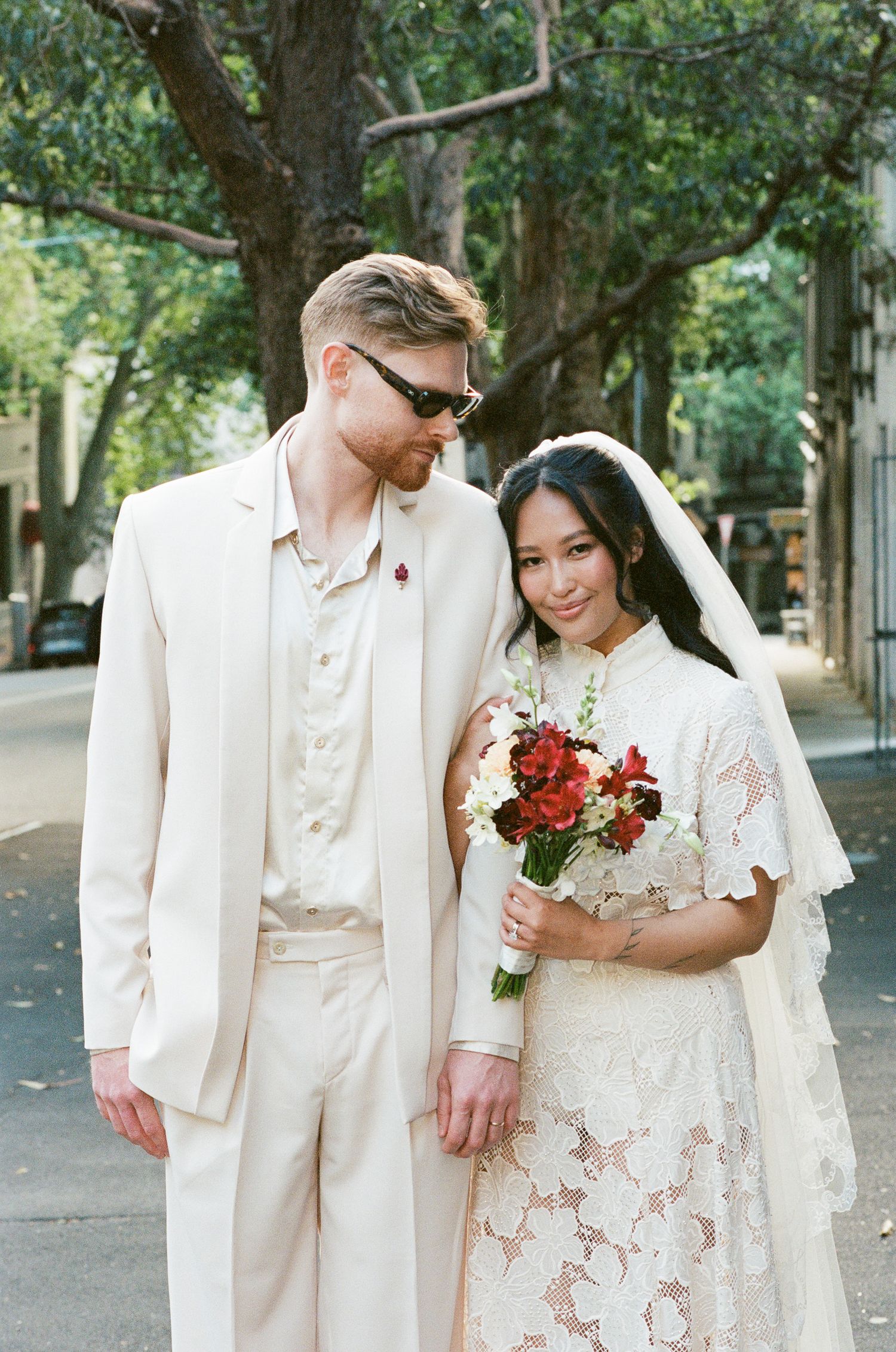 Bride in a lace dress and veil holds a red bouquet beside a groom in a cream suit and sunglasses on a tree-lined street.