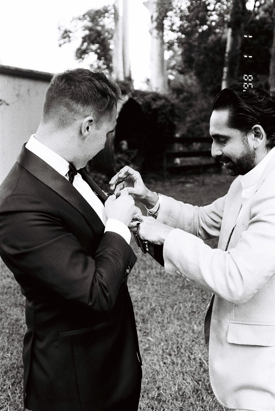 Black and white photo of two grooms in suits helping each other with boutonnieres before wedding ceremony.