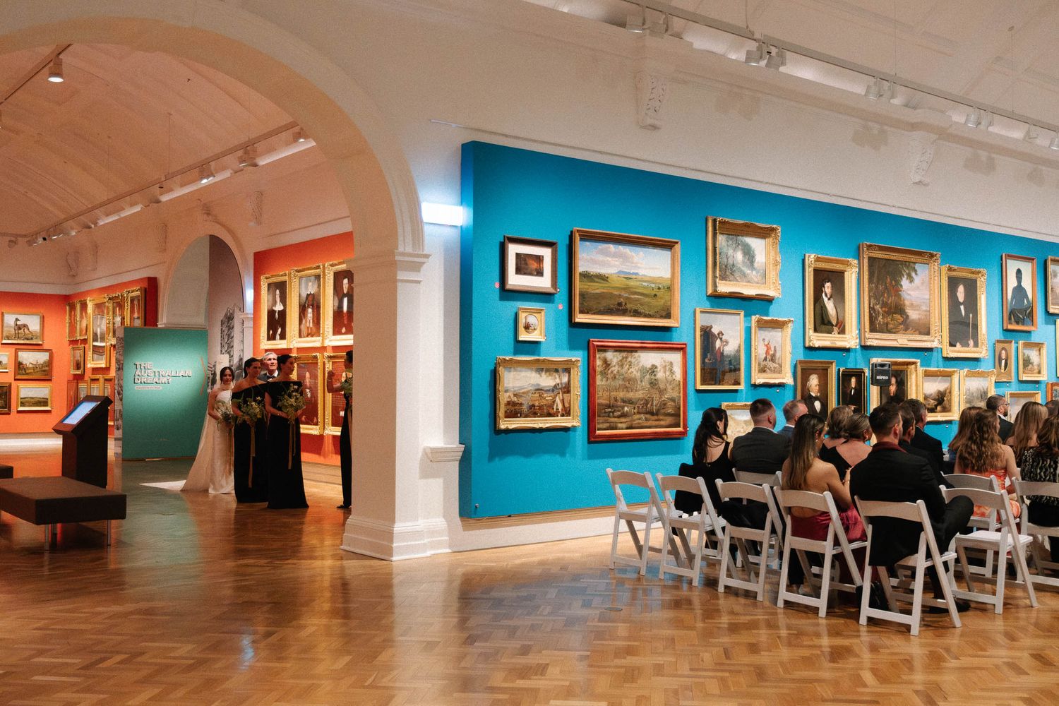 Art gallery interior with turquoise walls displaying framed paintings and chairs arranged for viewing in a spacious exhibition space.
