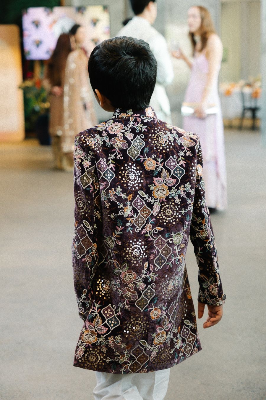 A person wearing an ornate floral and geometric patterned jacket walks away from the camera in a hallway.