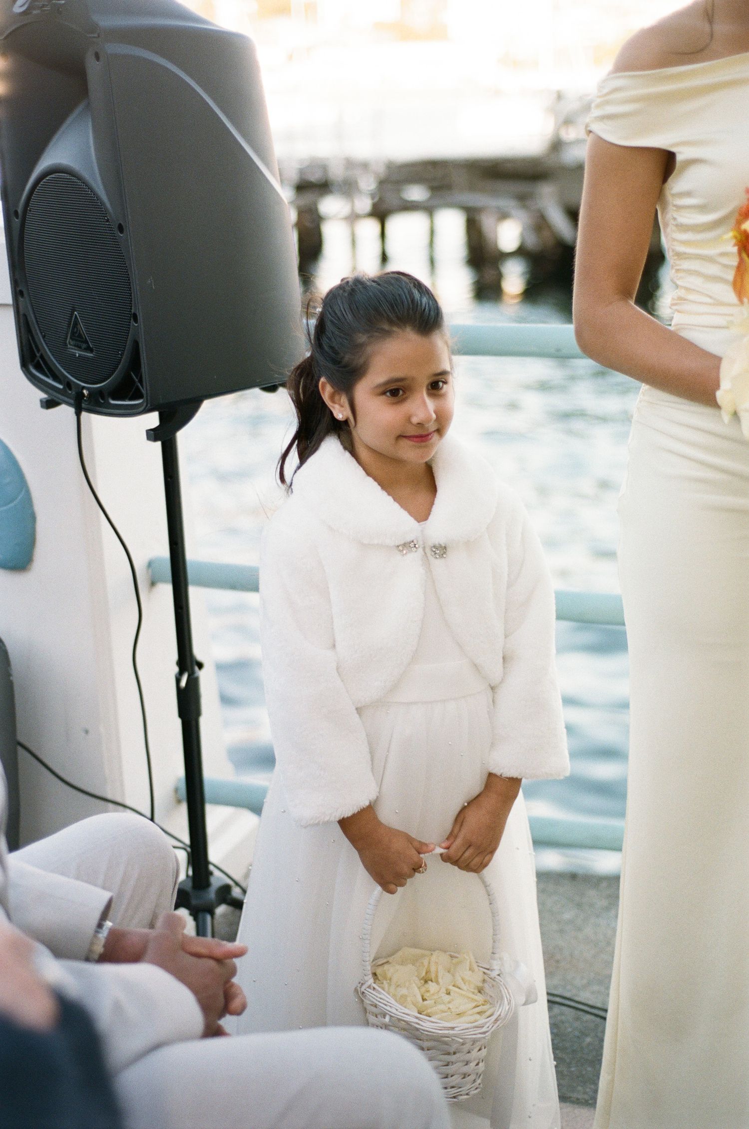 Young flower girl in a white dress and fur shawl holds a basket of petals at a waterfront wedding ceremony beside a woman in a cream gown.