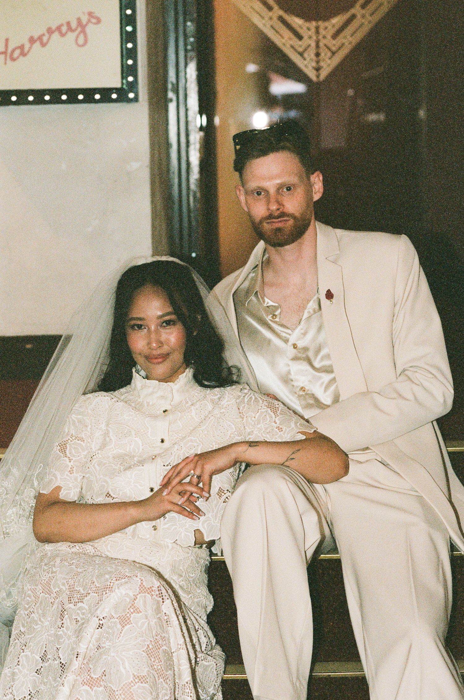 A bride in a white lace dress and veil sits beside a groom in a cream suit with a red boutonniere on vintage theater steps.