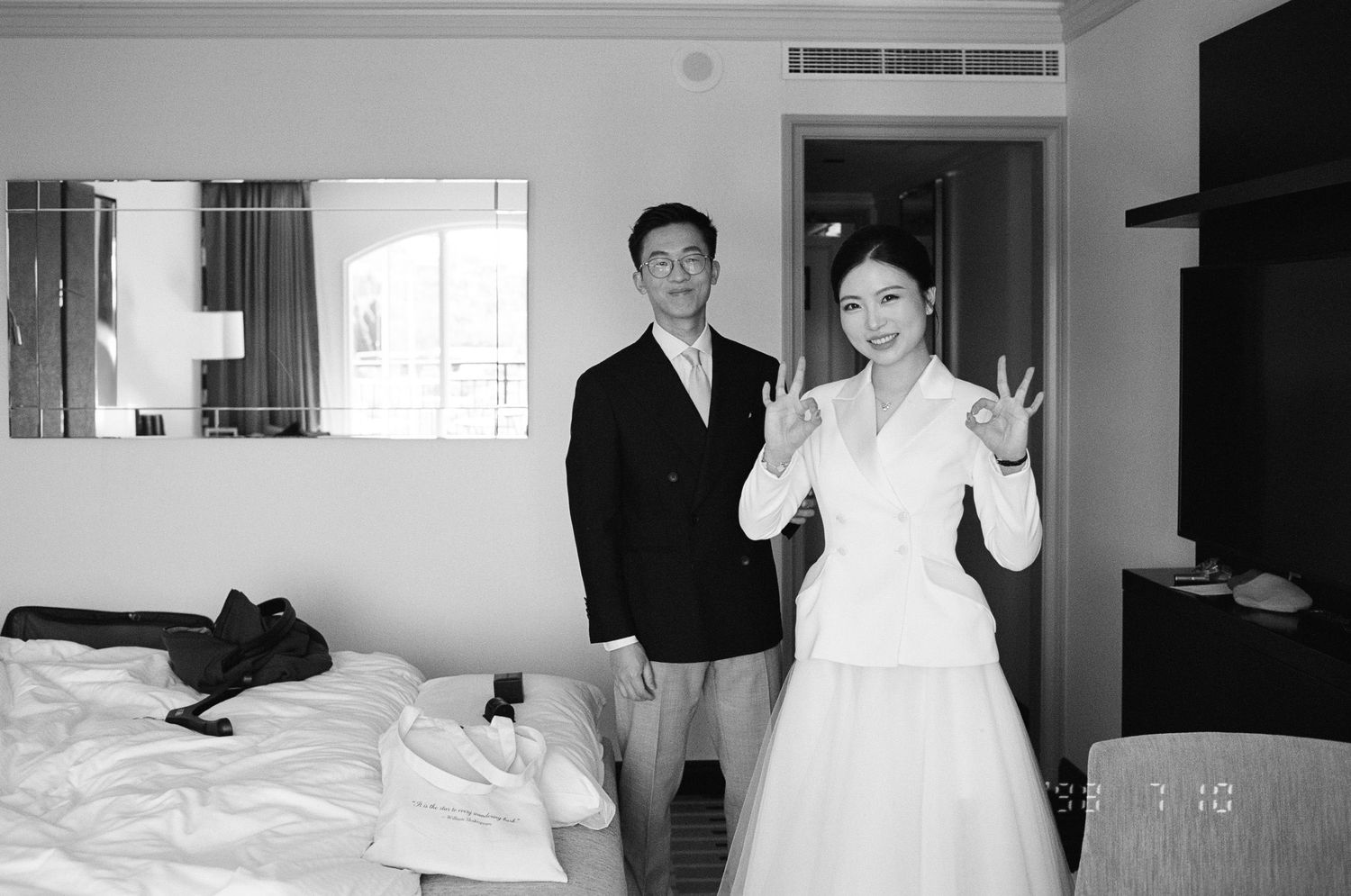 Black and white photo of a smiling couple in a hotel room, the woman in a white dress making peace signs beside a man in a dark suit.