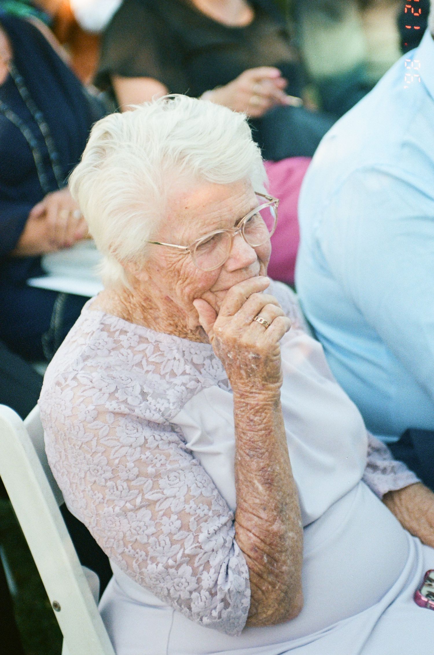 Elderly woman with white hair and glasses rests her hand on her chin, watching thoughtfully during an outdoor event with seated guests.
