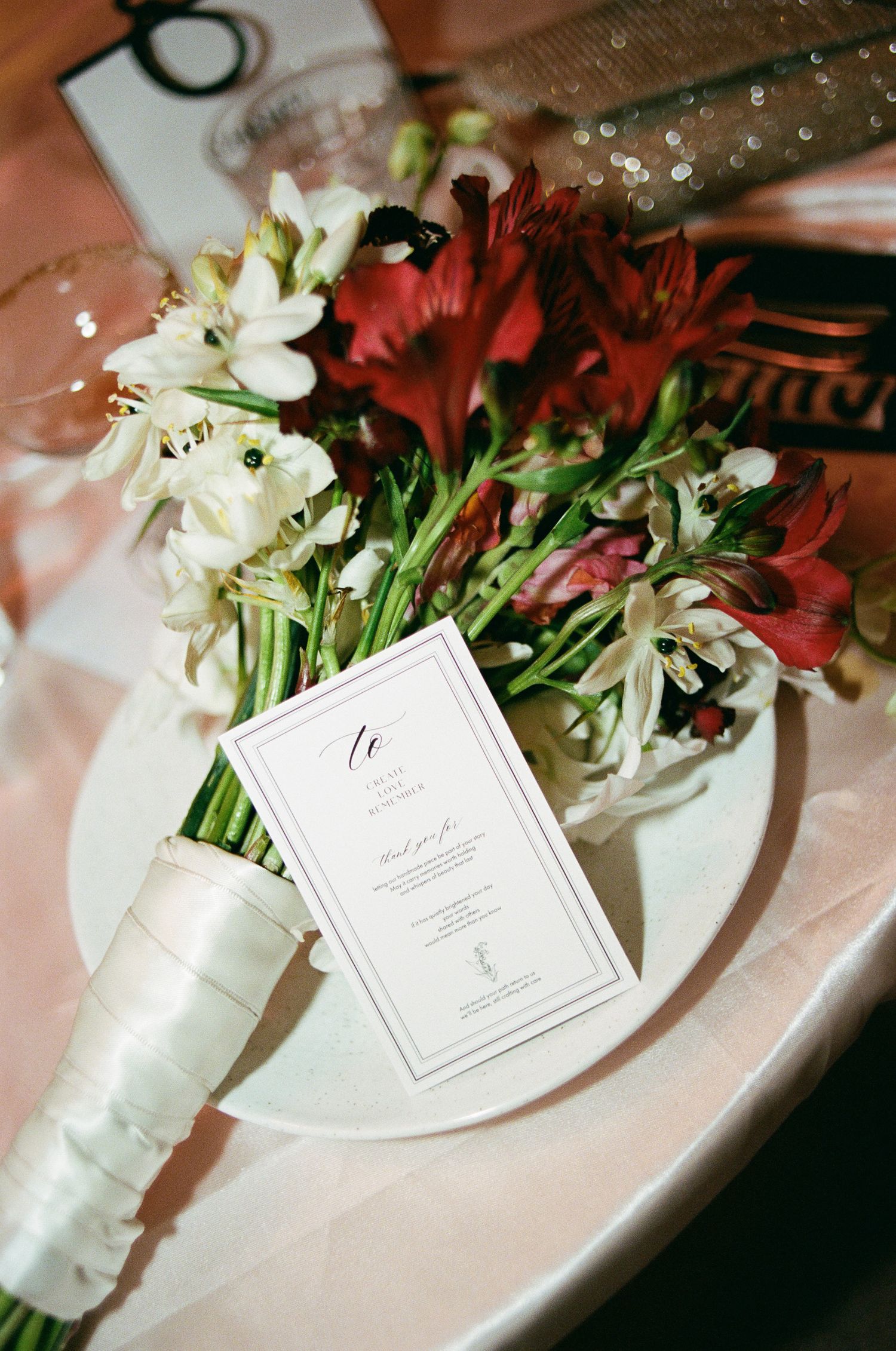 Wedding bouquet of red and white alstroemeria wrapped in ivory satin ribbon rests on a plate with an elegant printed menu card.