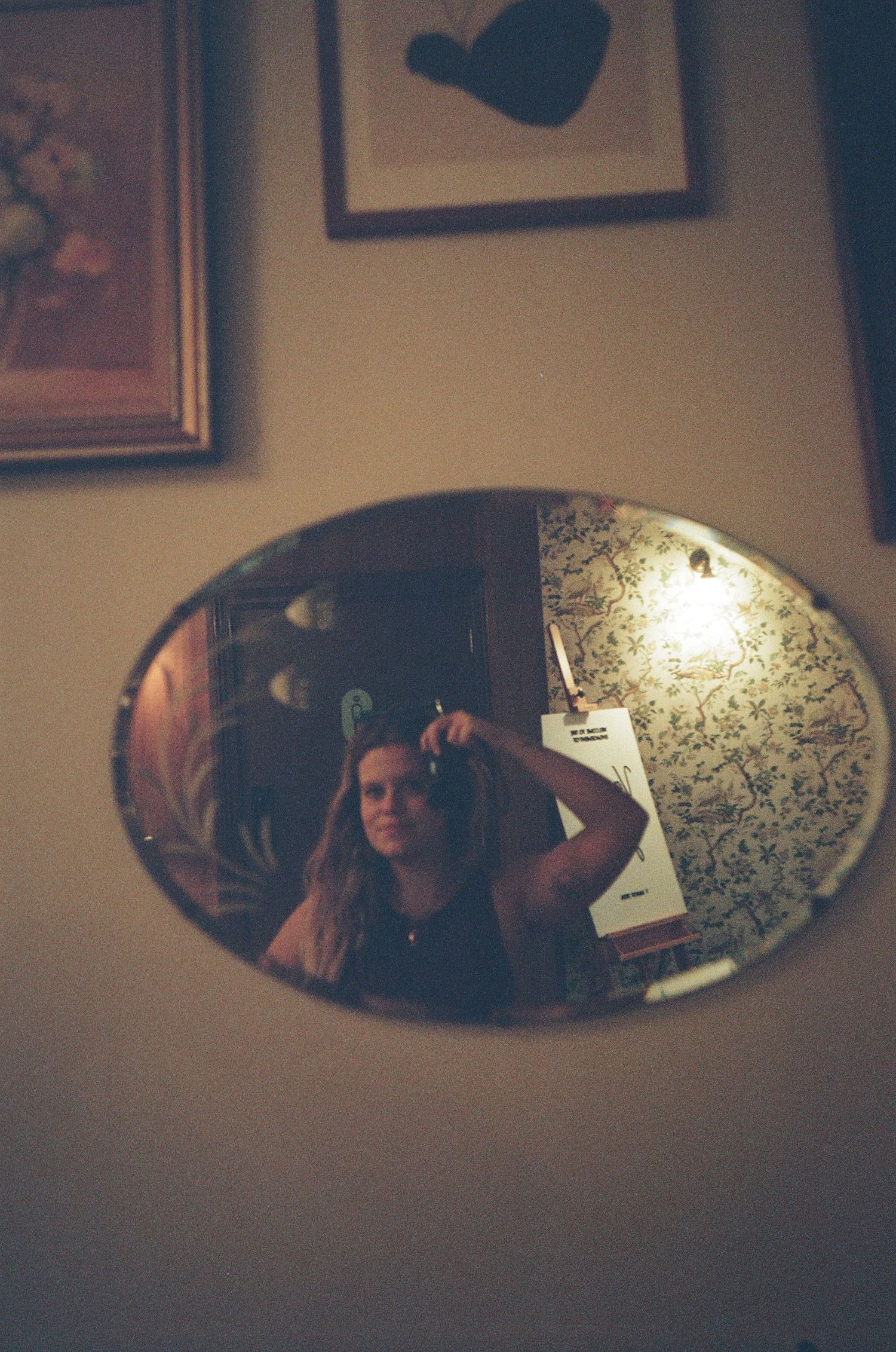 A young woman takes a selfie in an oval mirror on a wall, surrounded by framed artwork in a dimly lit room.