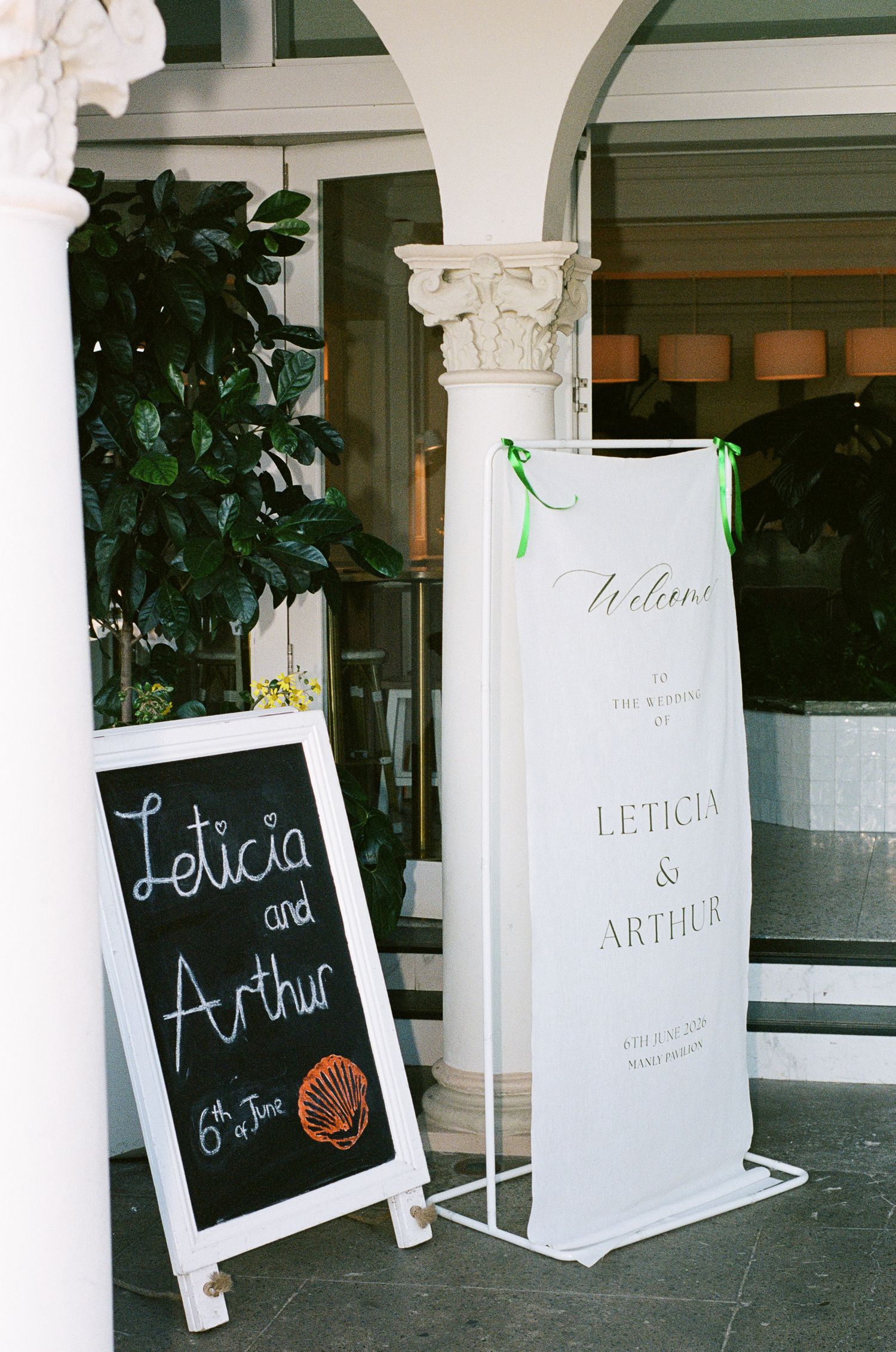 Wedding welcome sign for Leticia and Arthur beside a chalkboard with a shell drawing, set between white columns at an elegant entrance.
