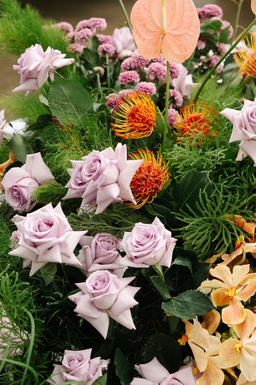 A stunning floral arrangement featuring lavender roses, orange pin cushion proteas, and delicate green foliage.