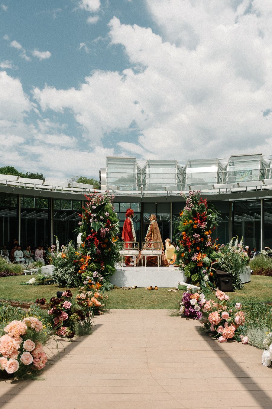 Elegant outdoor wedding ceremony setup with floral arches and a paved aisle leading to a modern glass building backdrop.