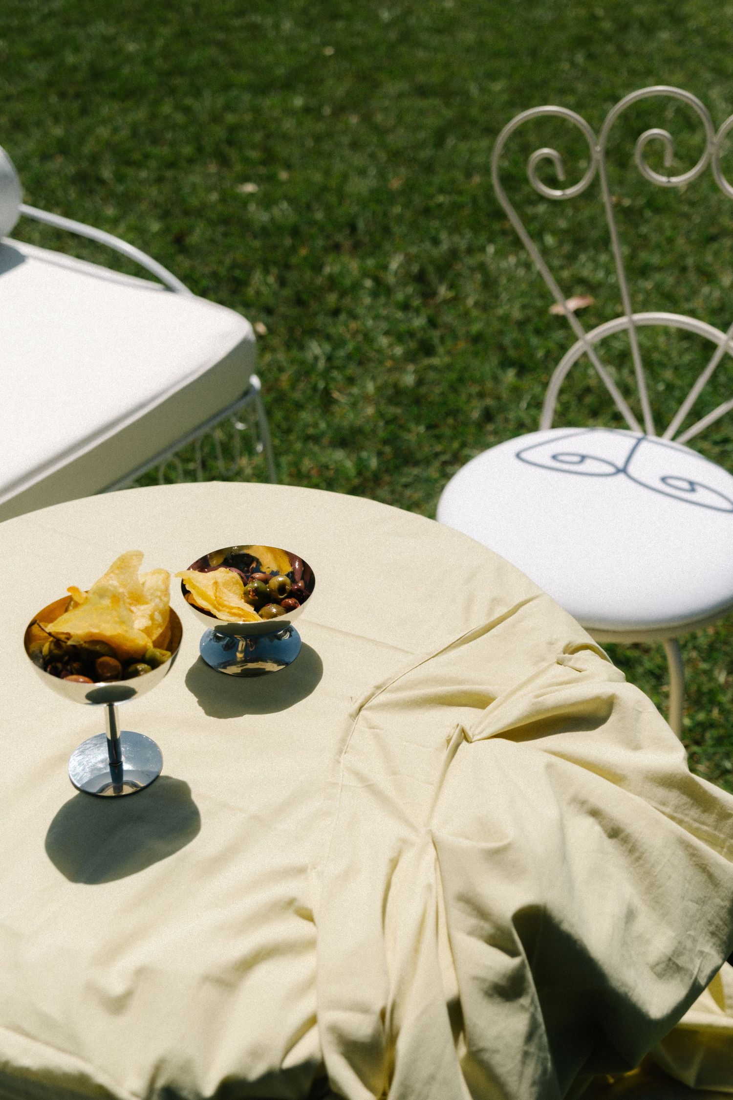 Two cocktail glasses on a cream tablecloth with ornate white garden chair in an outdoor setting.