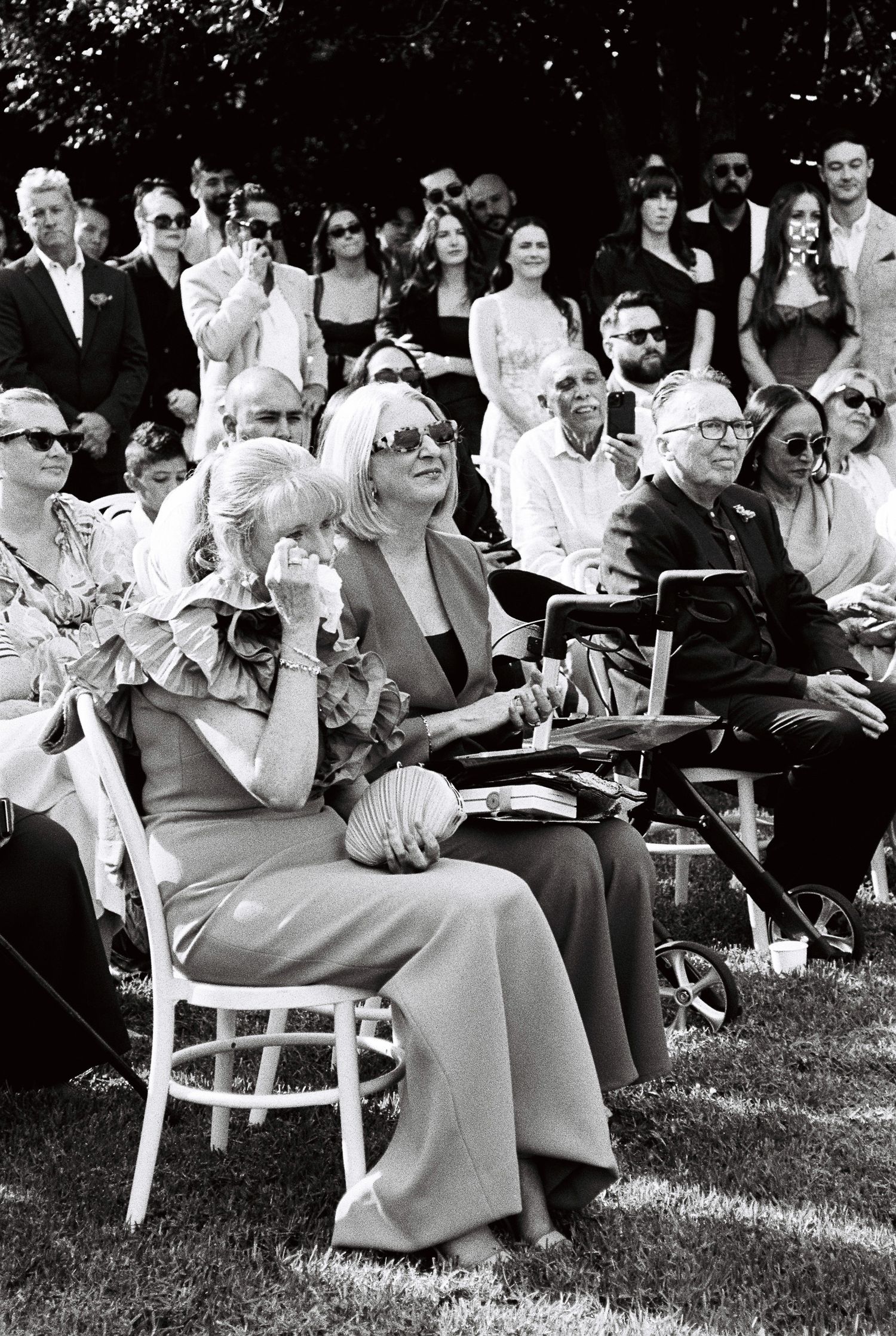Emotional wedding guests seated outdoors on white chairs, with a woman in a ruffled dress wiping a tear during the ceremony.