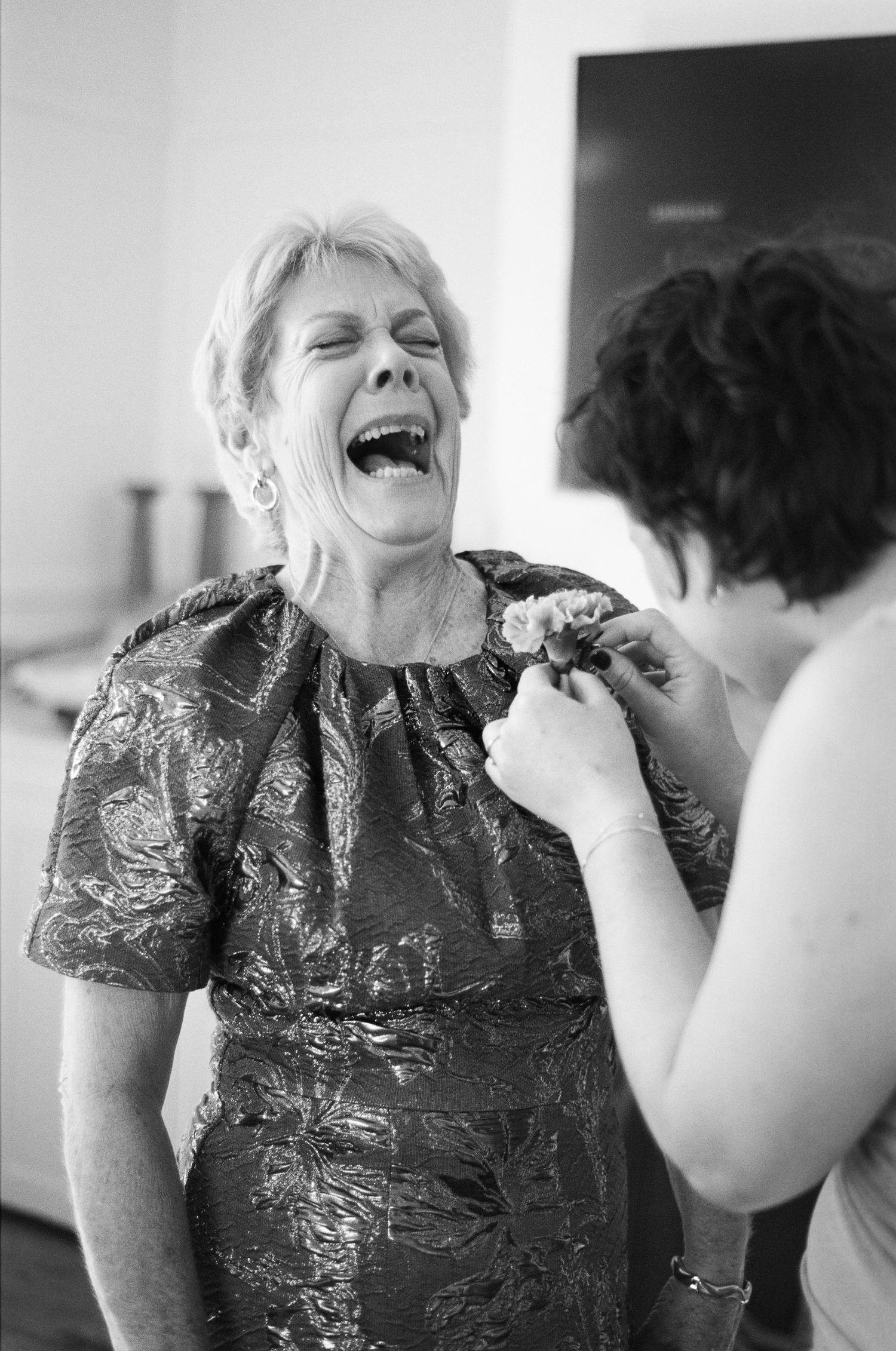 Black and white photo of a laughing older woman in a metallic brocade dress as another woman pins a flower boutonniere to her.