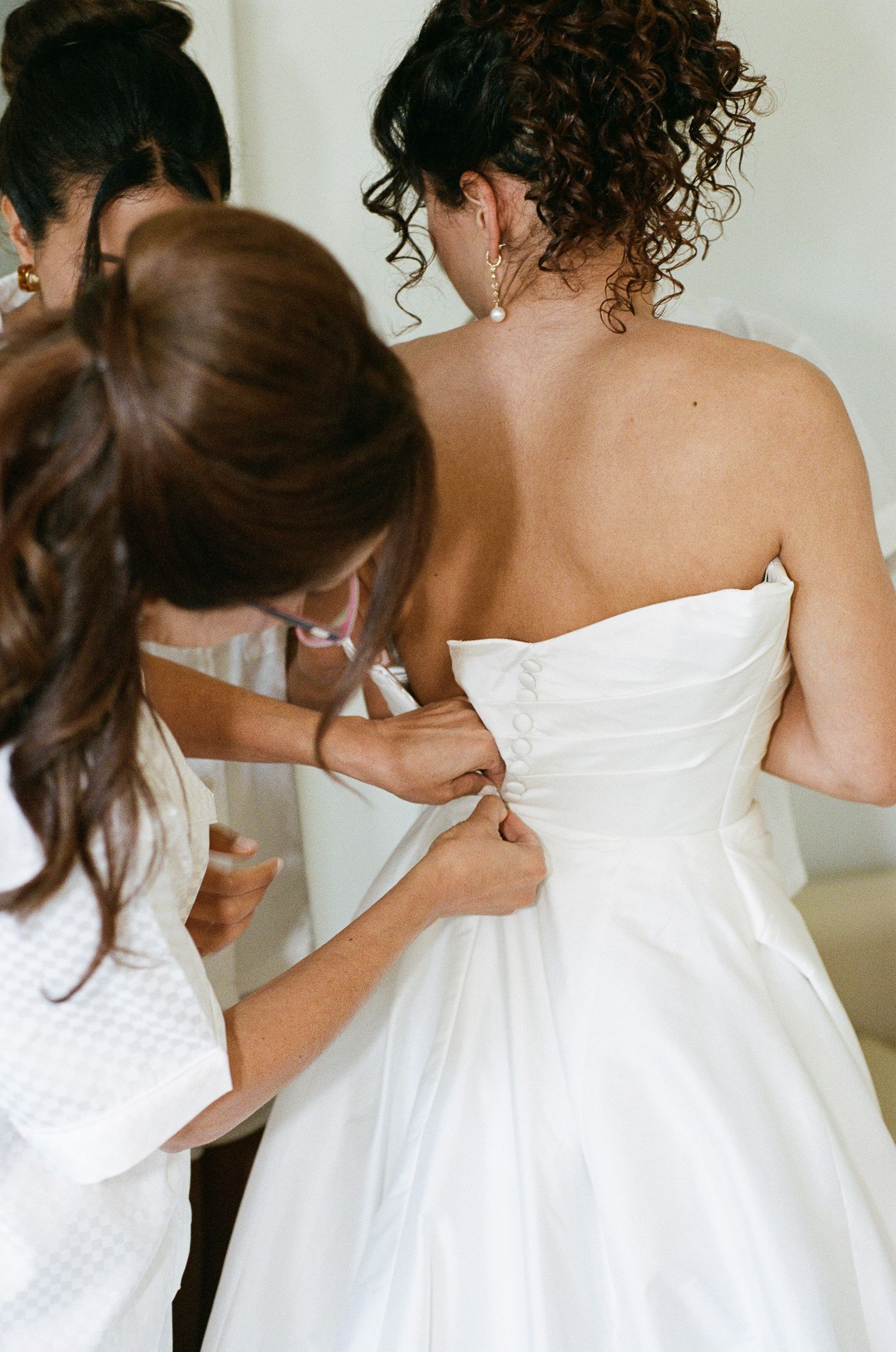 Bridesmaids in white robes help button the back of a bride's strapless white wedding gown before the ceremony.