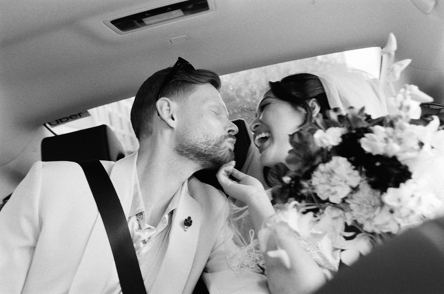 Black and white photo of a smiling bride holding a bouquet and a groom in a light suit leaning in for a kiss inside a car.