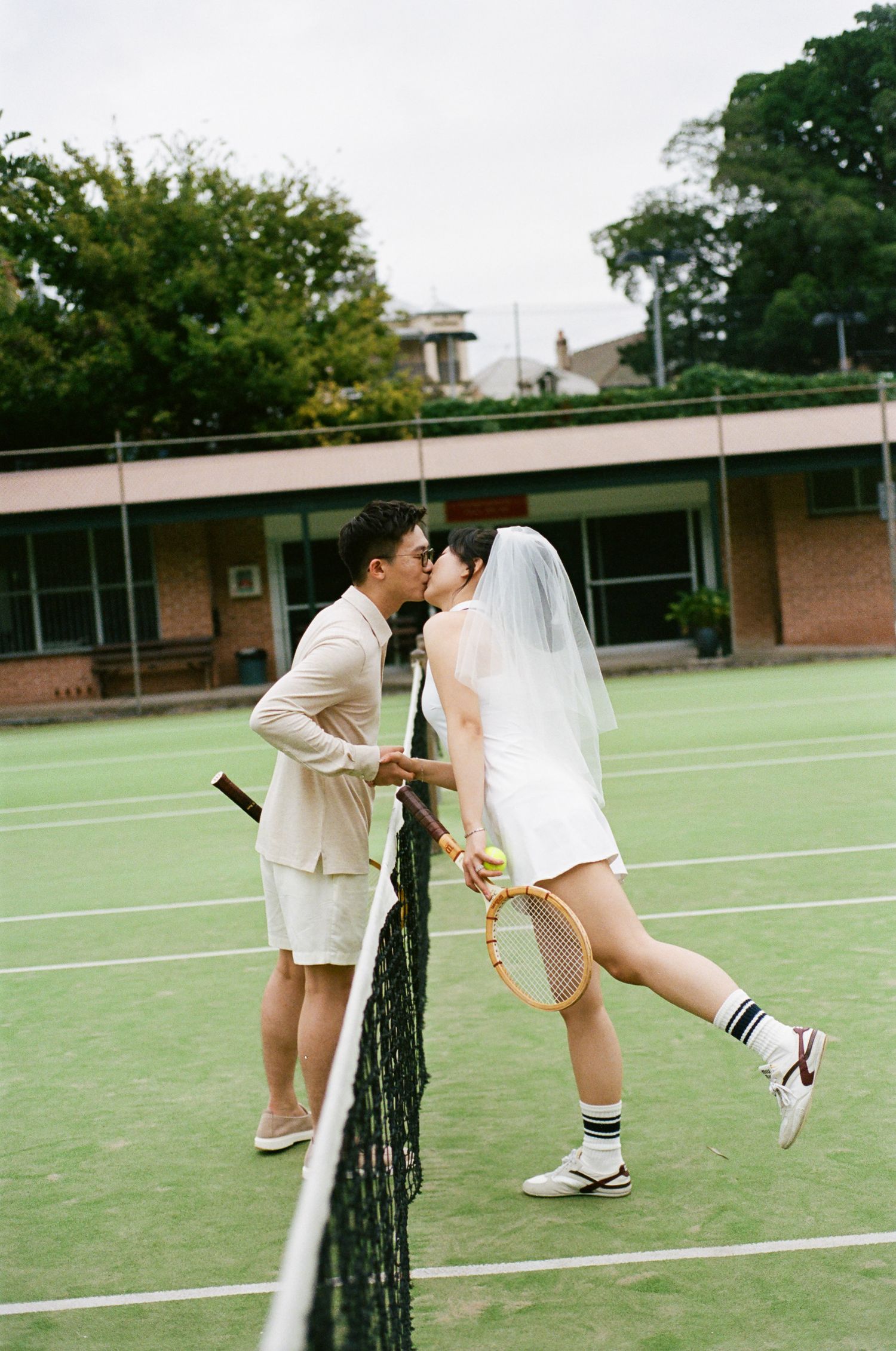 Couple in tennis attire kiss over a tennis court net, the bride wearing a veil and holding a racket, with a brick clubhouse behind.