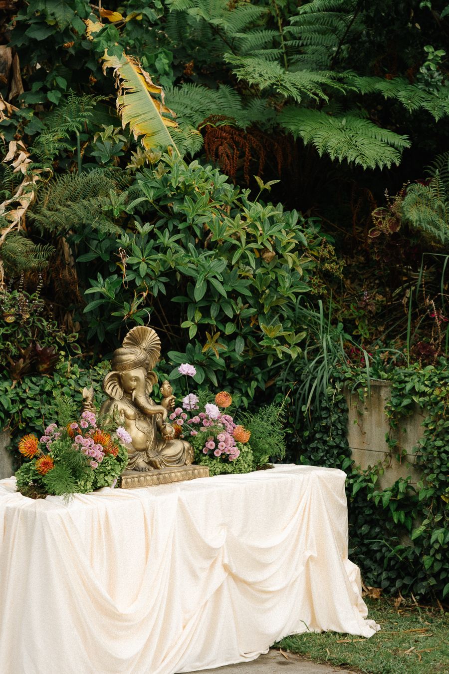 An elegant white draped table with a stone statue and colorful floral arrangements set against lush garden greenery.