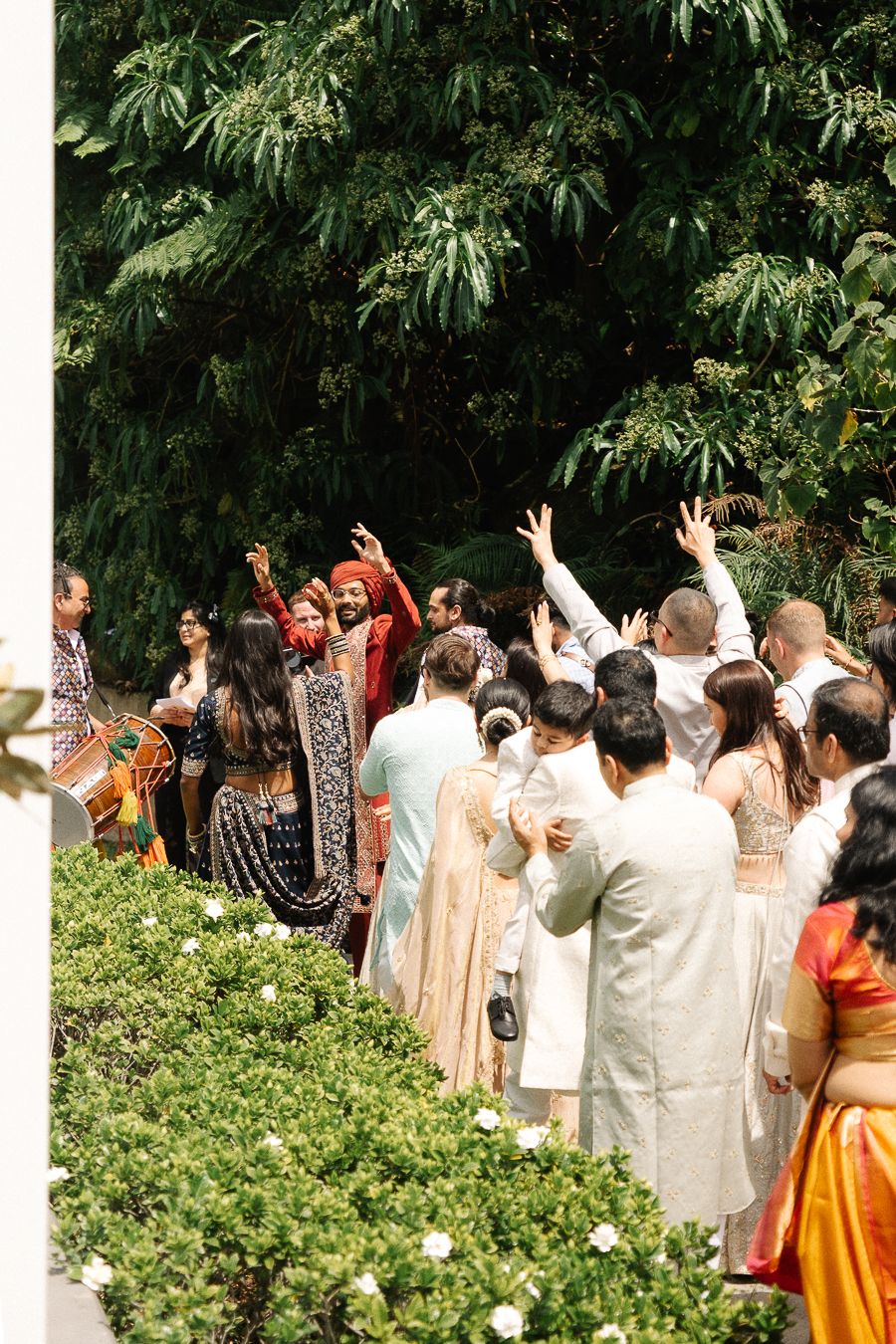 A group of wedding guests in formal attire raise their hands in celebration outdoors near lush greenery.