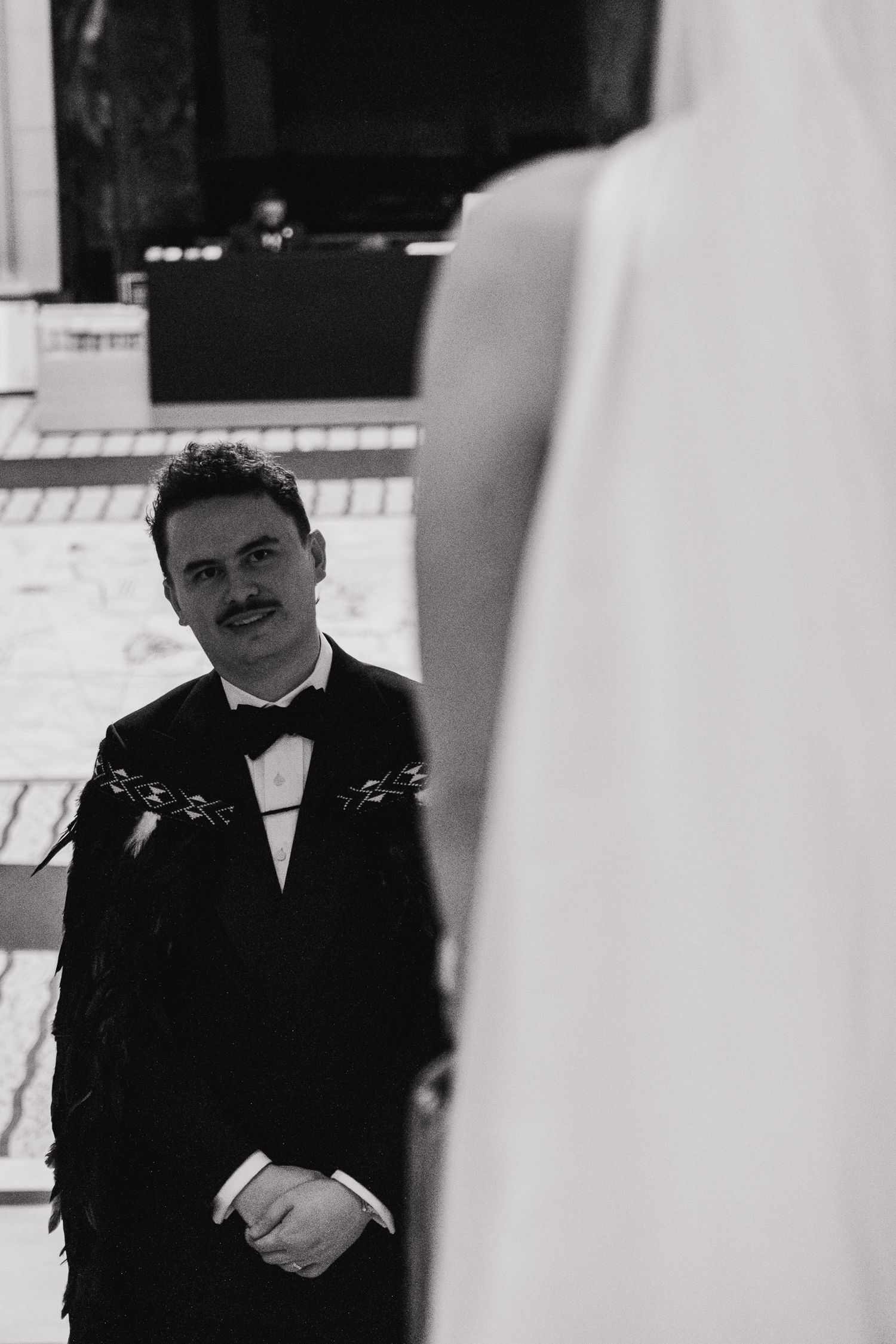 A person in a black tuxedo and bow tie stands near white columns in a formal setting.