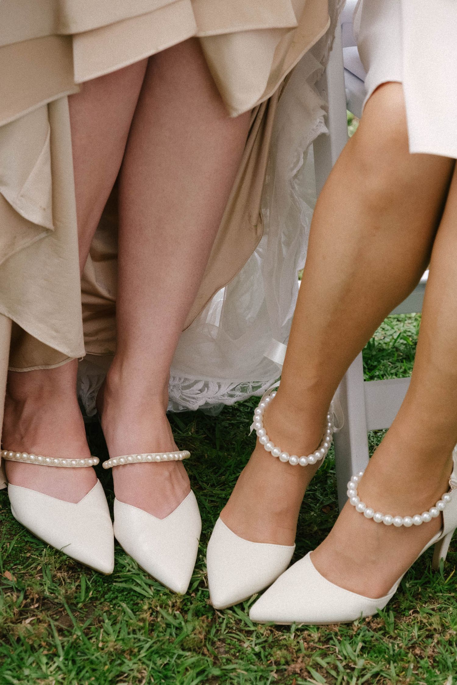 Two pairs of elegant white bridal shoes with pearl ankle straps shown on grass at a wedding.