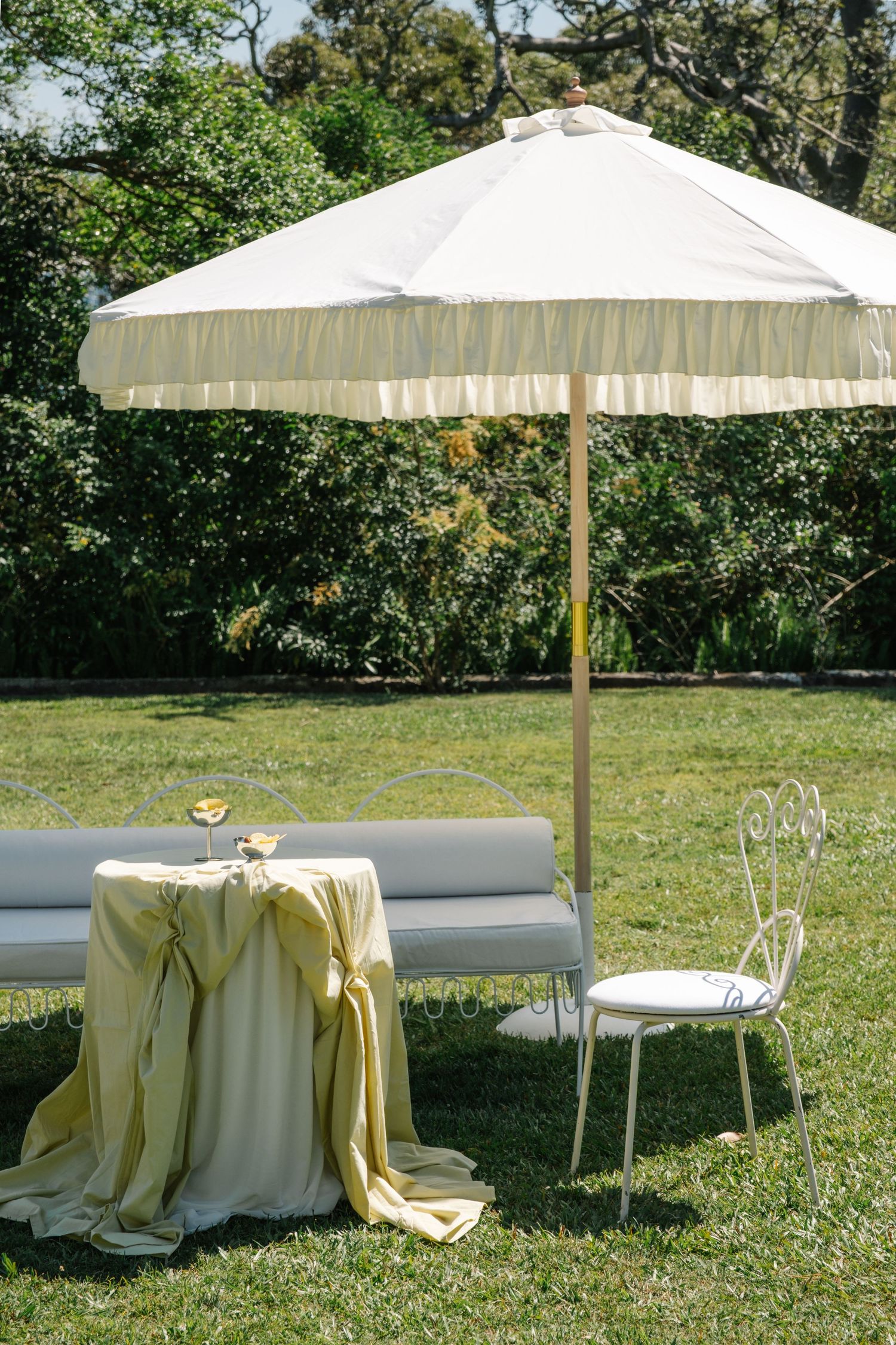 Elegant garden setup with white umbrella, draped table and metal chair on green lawn surrounded by trees.