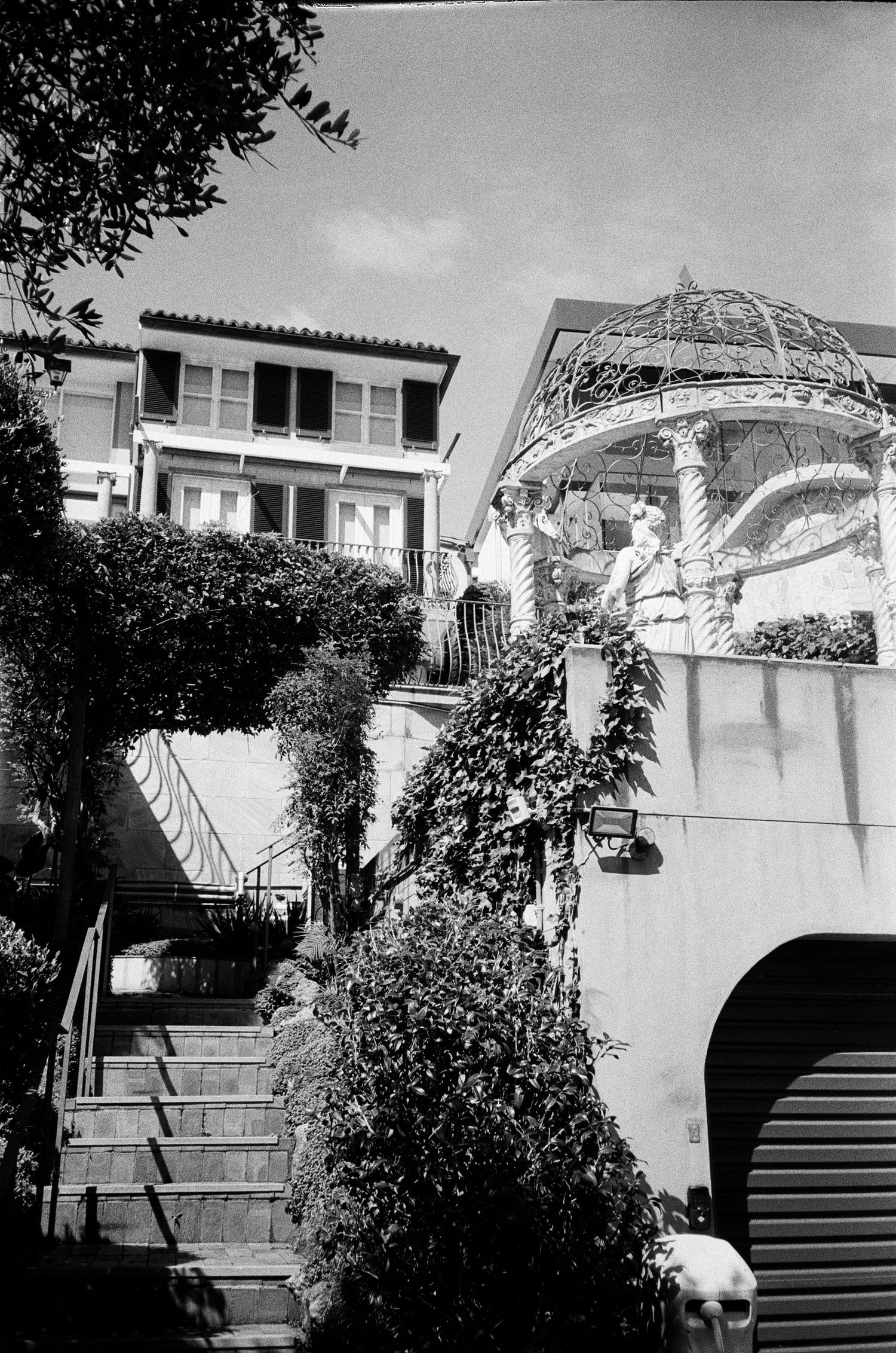 Black and white photographs showing a decorative metal gazebo structure on top of a stairway entrance to a house.