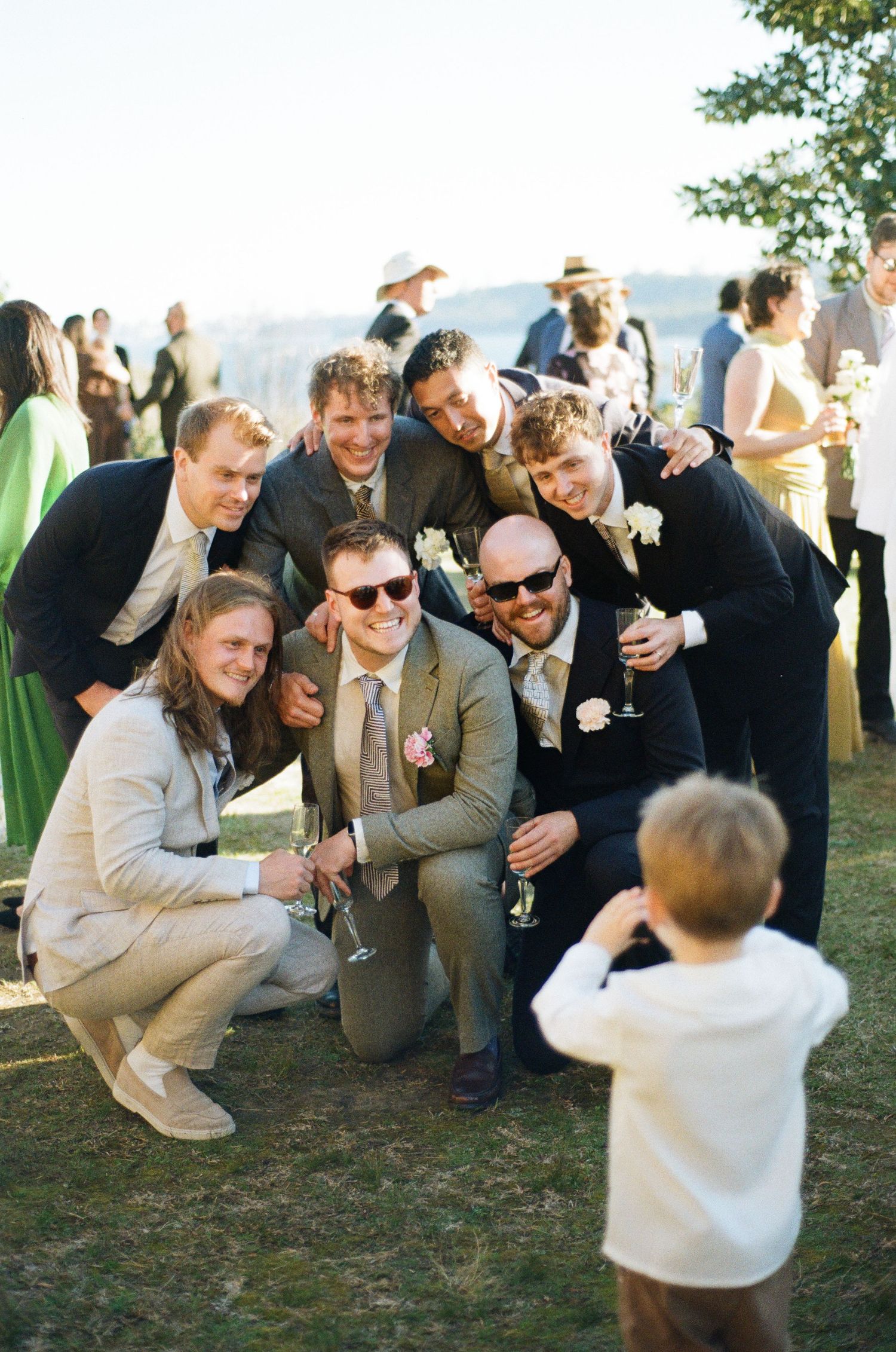 Groomsmen posing for a photo with a young boy at an outdoor wedding reception on a sunny day.