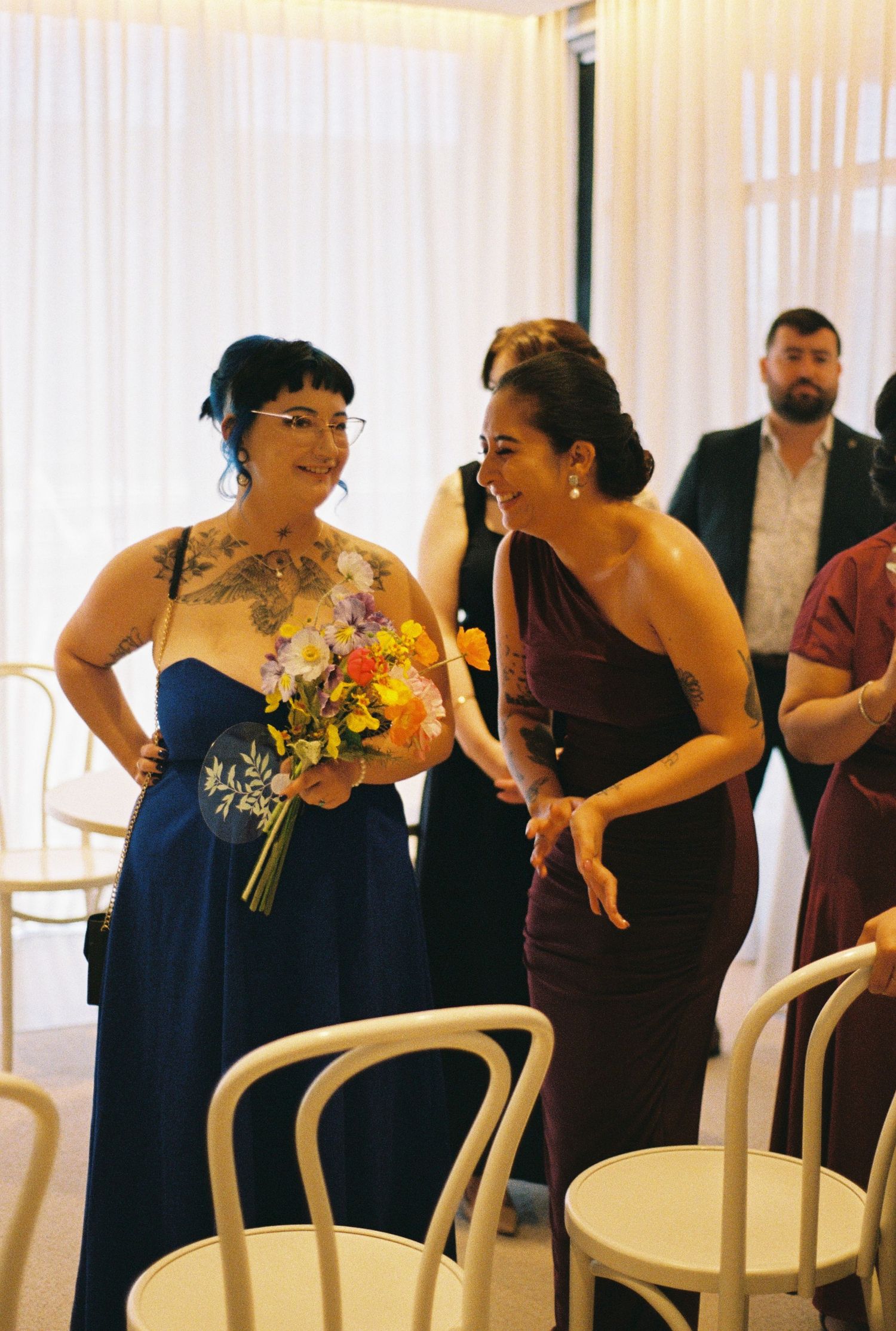 Two people in formal attire share a moment at a wedding ceremony with white chairs and sheer curtains in the background.