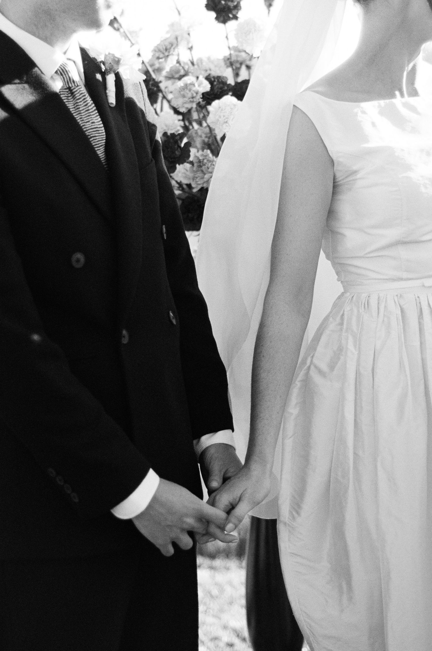 Black and white photo of a bride in a sleeveless gown and veil holding hands with a groom in a suit during a wedding ceremony.
