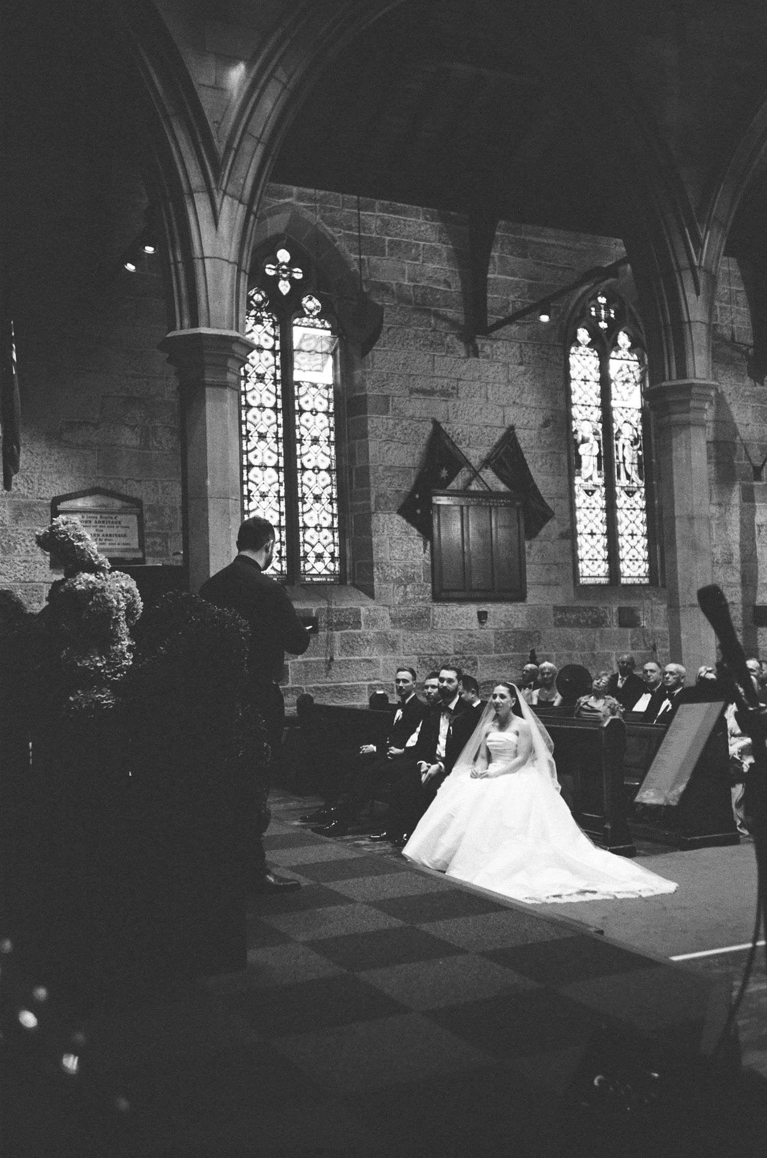 Black and white photo of a bride in a white gown seated in a stone church with stained glass windows as wedding guests look on.
