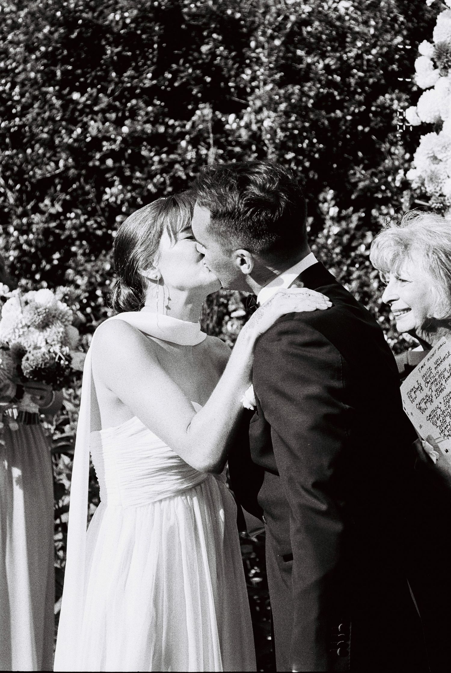 Black and white photo of a bride and groom sharing their first kiss at an outdoor wedding ceremony as a smiling guest looks on.