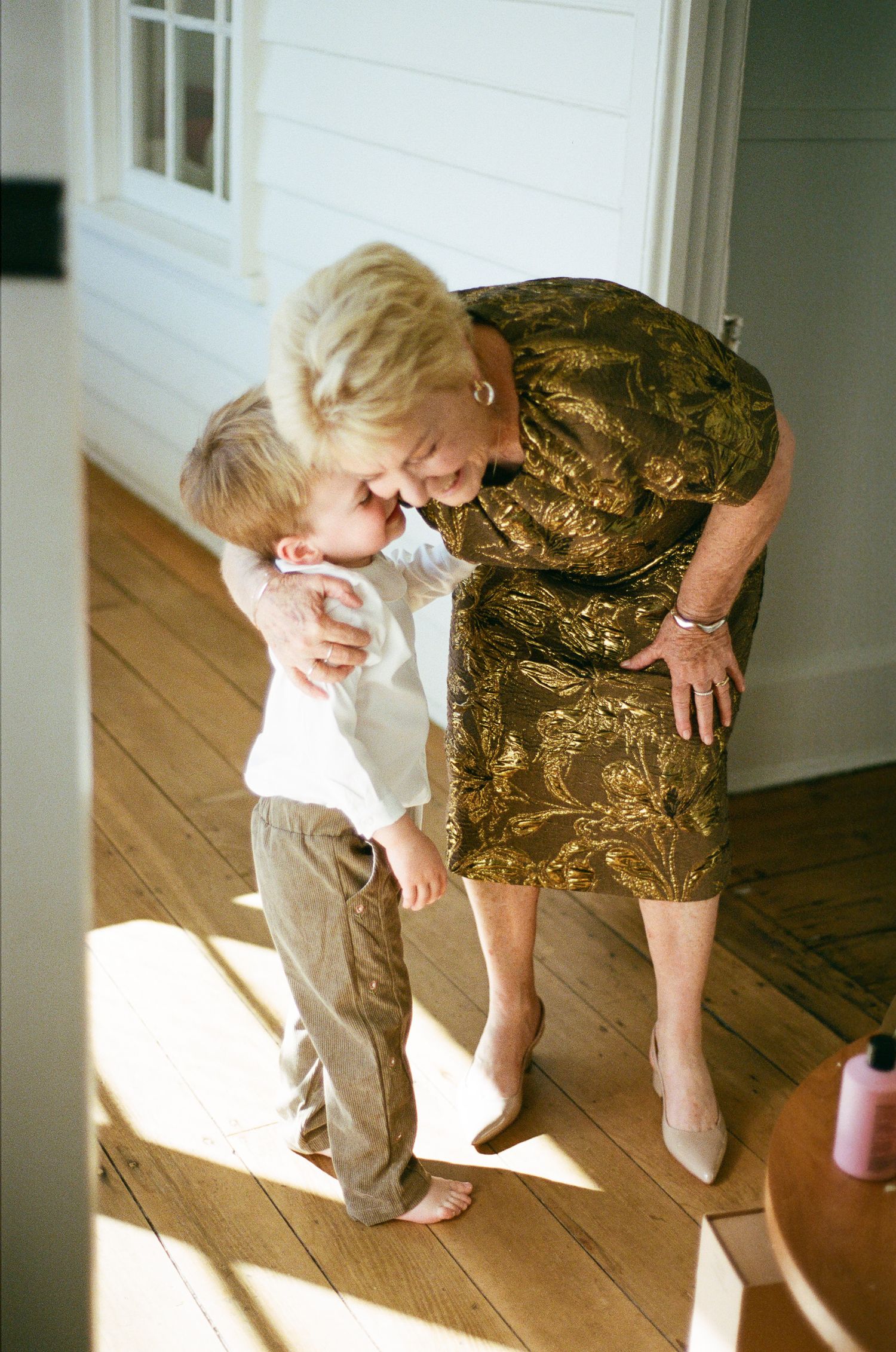 Smiling woman in a gold brocade dress bends down to hug a barefoot toddler in sunlit hardwood hallway.