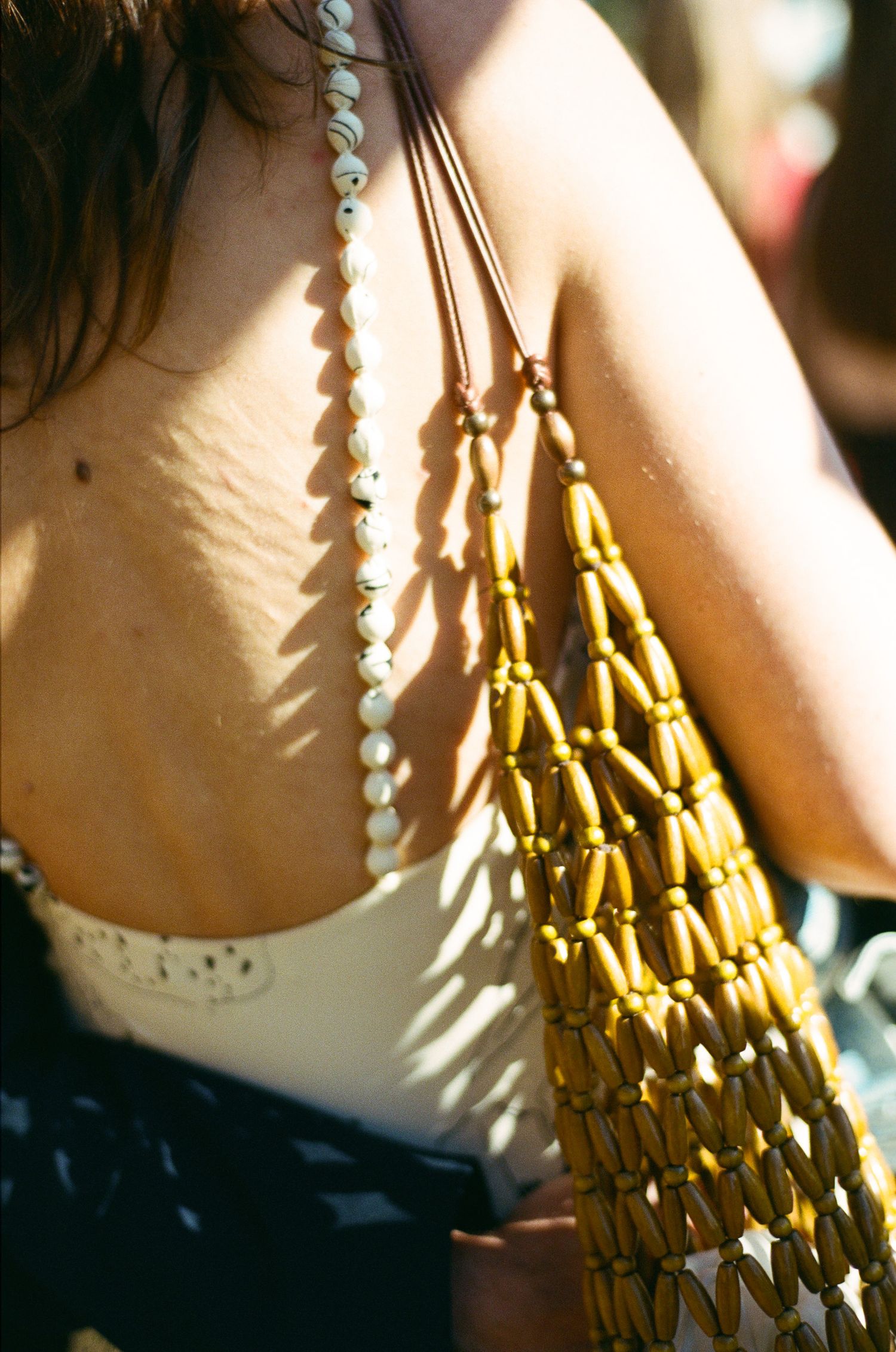 Woman's back in sunlight wearing a white top, with a beaded wooden bag and a white shell necklace hanging over her shoulder.