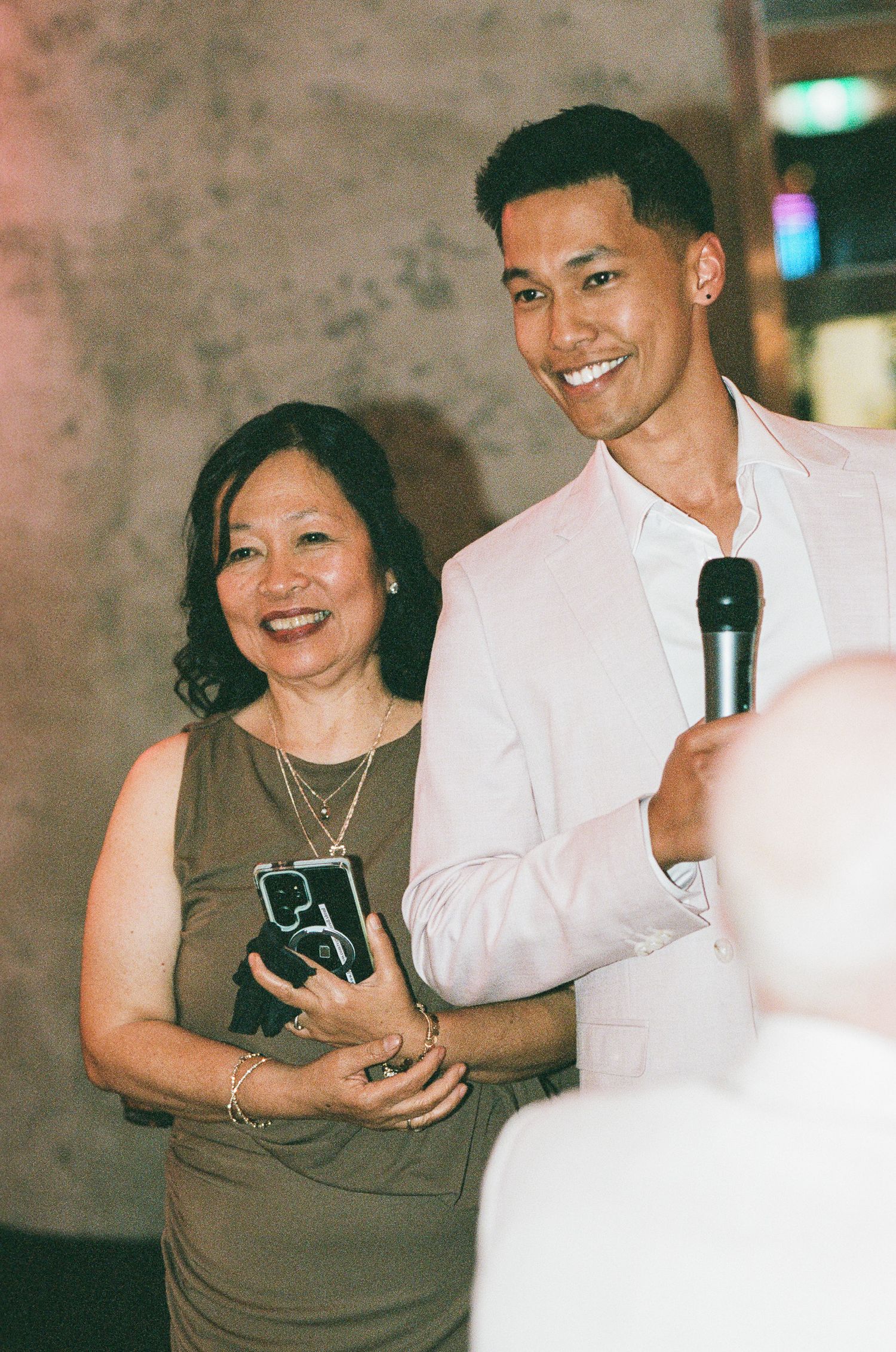 Smiling man in a light blazer holds a microphone beside a smiling woman in an olive dress holding a phone at an indoor event.