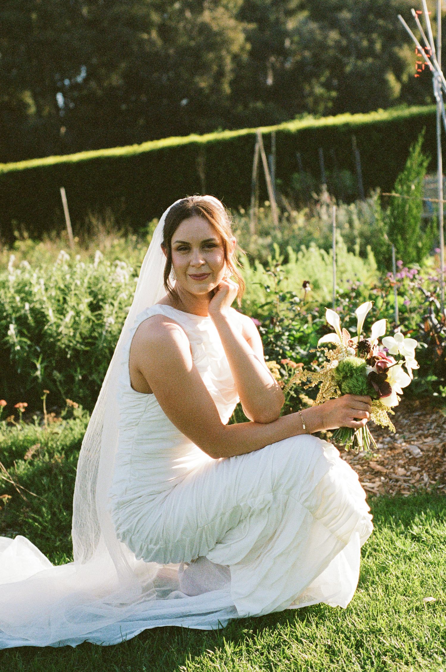 Smiling bride in a white gown and long veil crouches in a lush garden, holding a wildflower bouquet of white and pink blooms.