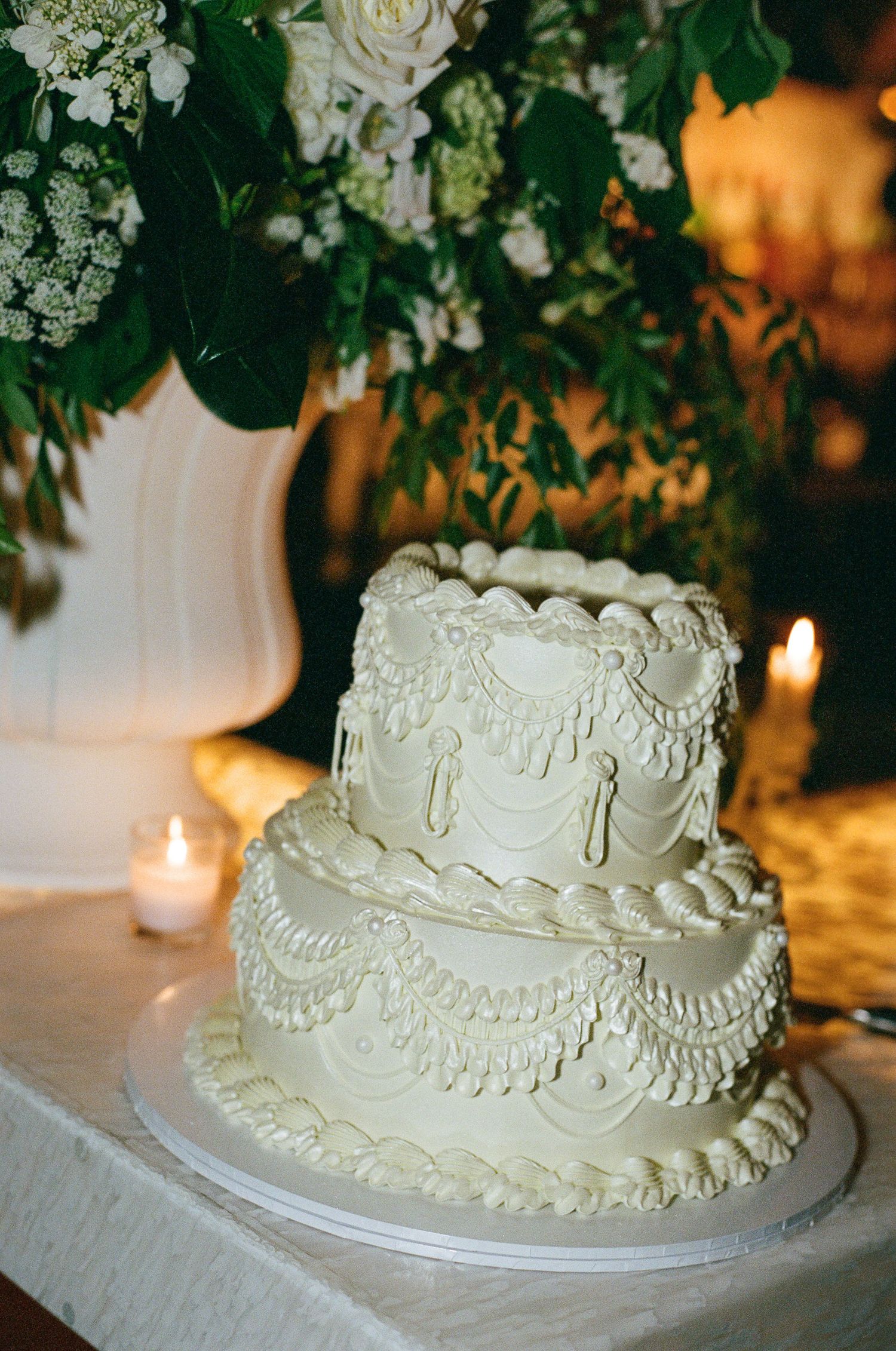 Two-tier white vintage-style wedding cake with piped lace frosting sits on a draped table beside white flowers and glowing candles.