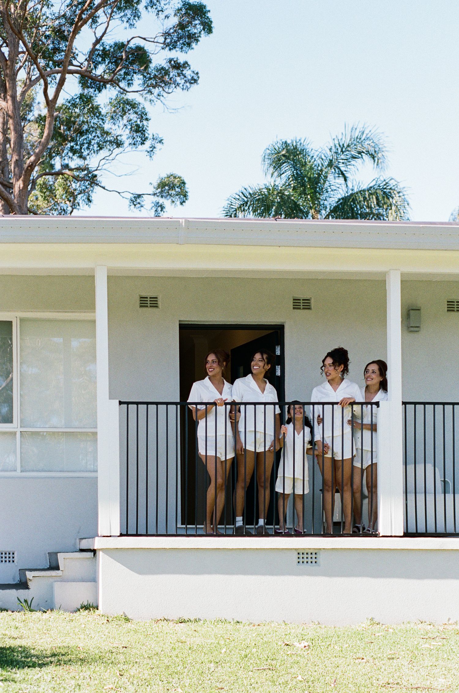 Five women in matching white robes smile and chat on the porch of a white single-story house with a green lawn and tall trees.