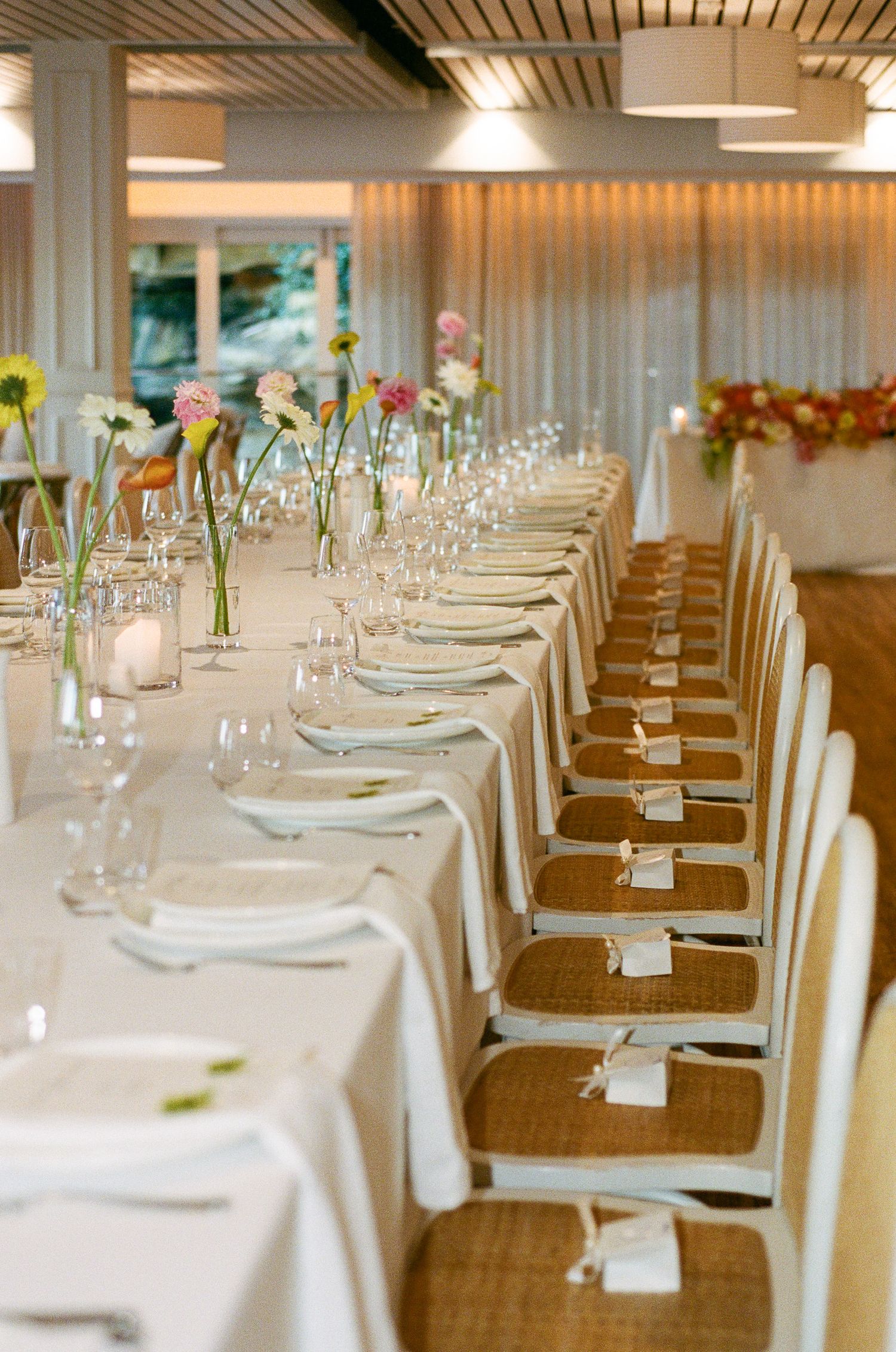 Long banquet table set with white linens, plates and glasses, with colorful gerbera daisies in vases and wicker chairs with place cards.