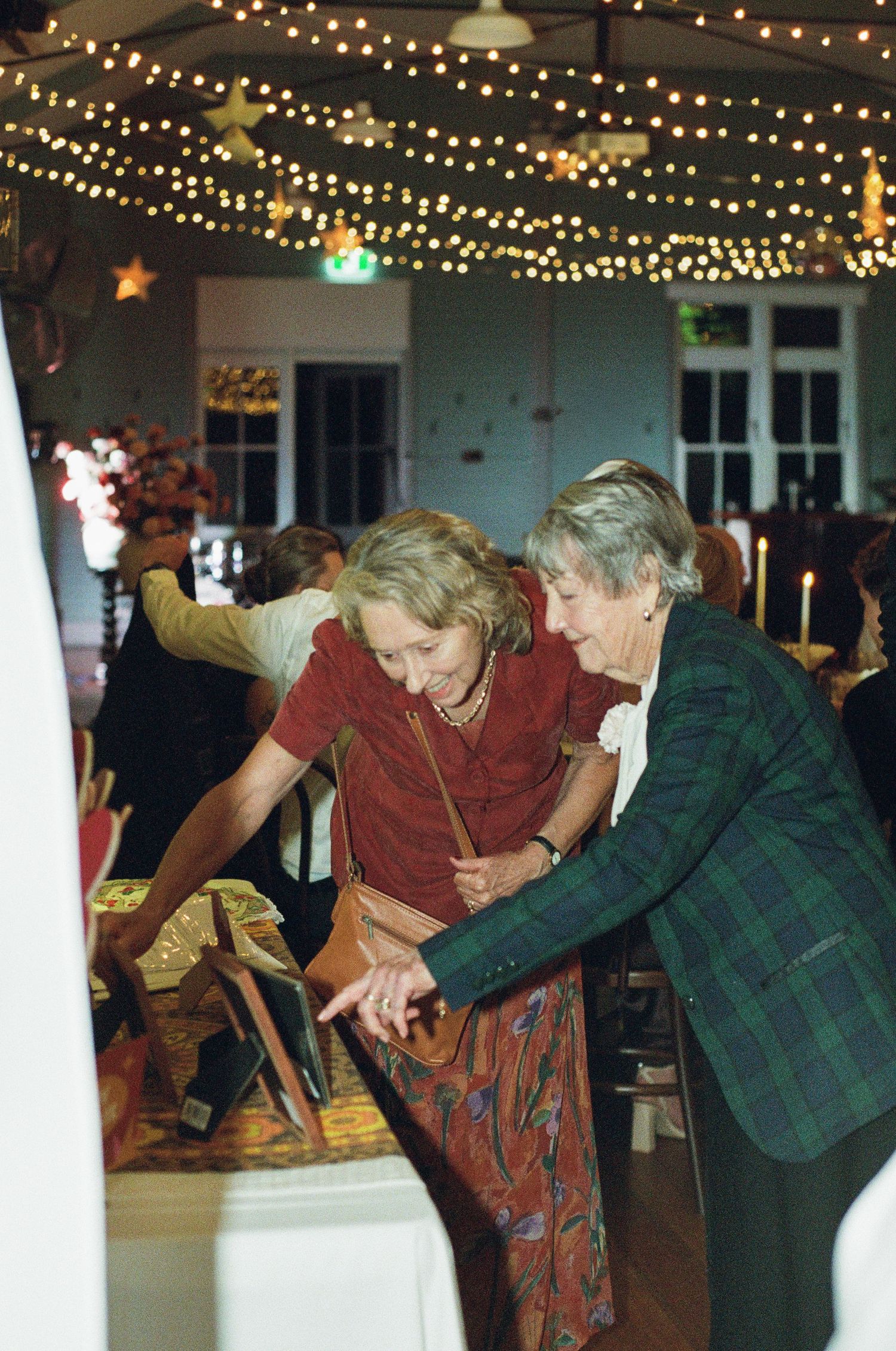 Two elderly women examining items together at decorated event venue with string lights overhead.