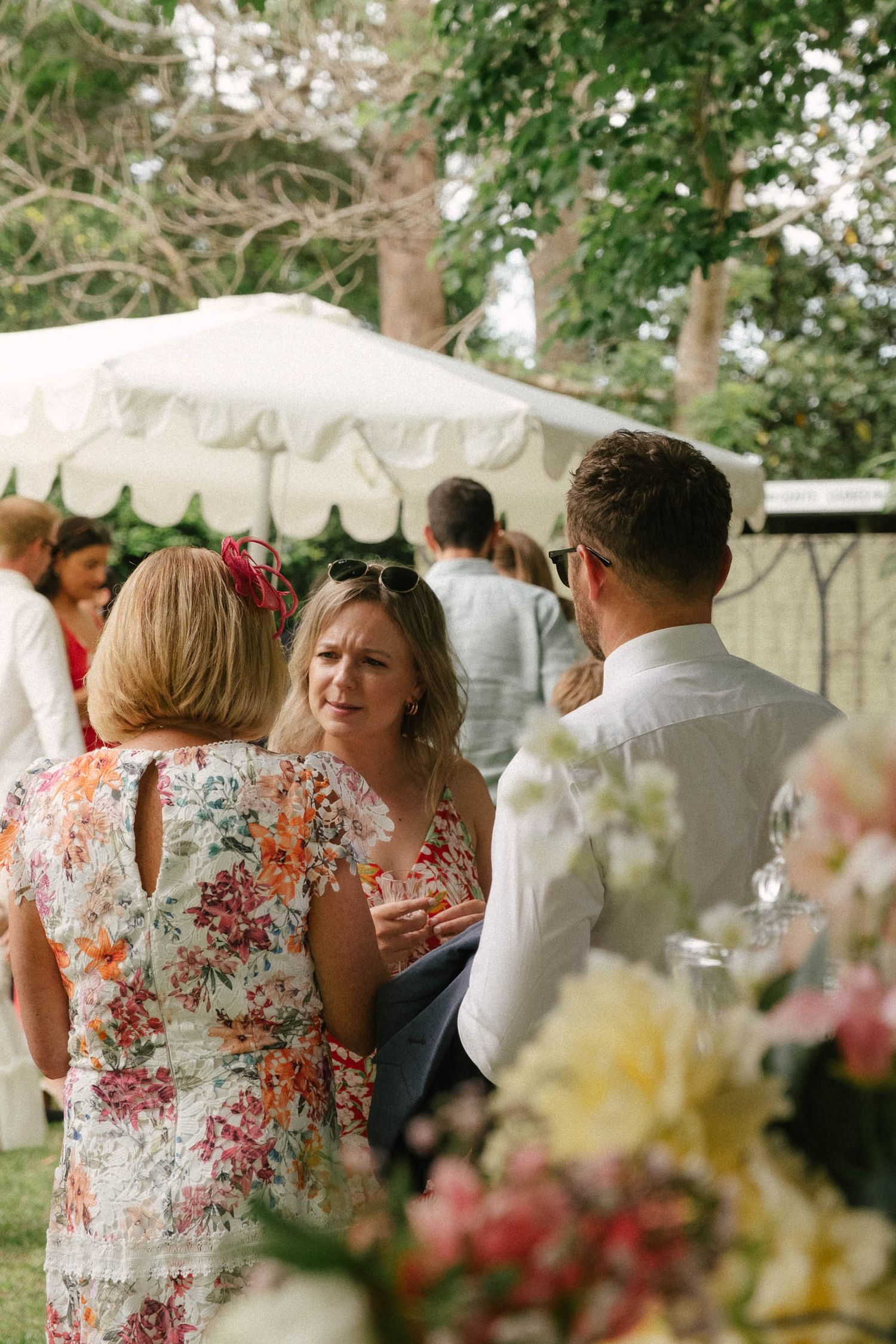 Guests mingle under a white tent canopy at an outdoor garden party with floral arrangements visible in foreground.