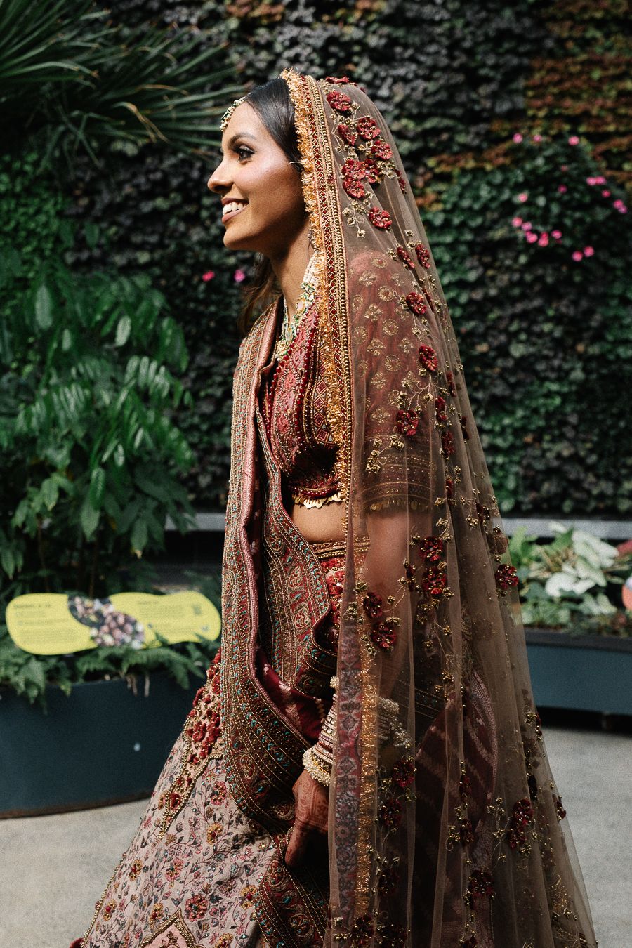 A bride in a traditional Indian wedding lehenga and veil poses elegantly in an outdoor garden setting.