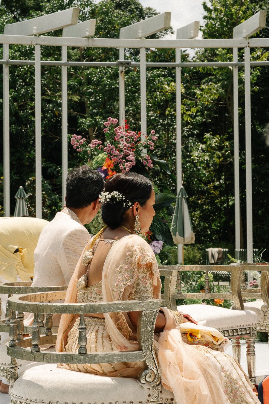 People wearing floral crowns and flowing dresses relax on elegant white outdoor furniture at a garden party.