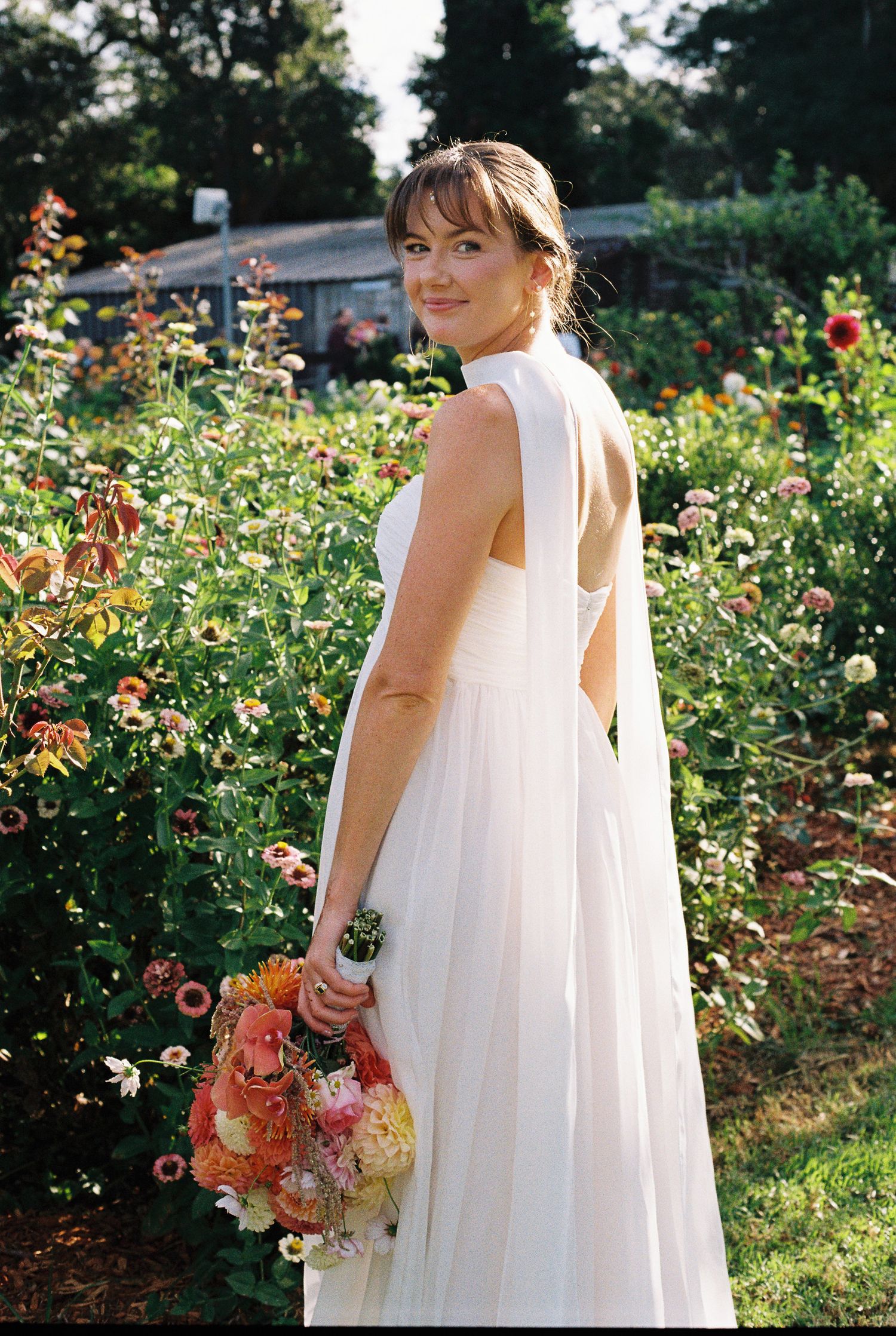 Bride in a flowing white gown with open back and sheer drape holds an orange and pink bouquet while standing in a blooming flower garden.
